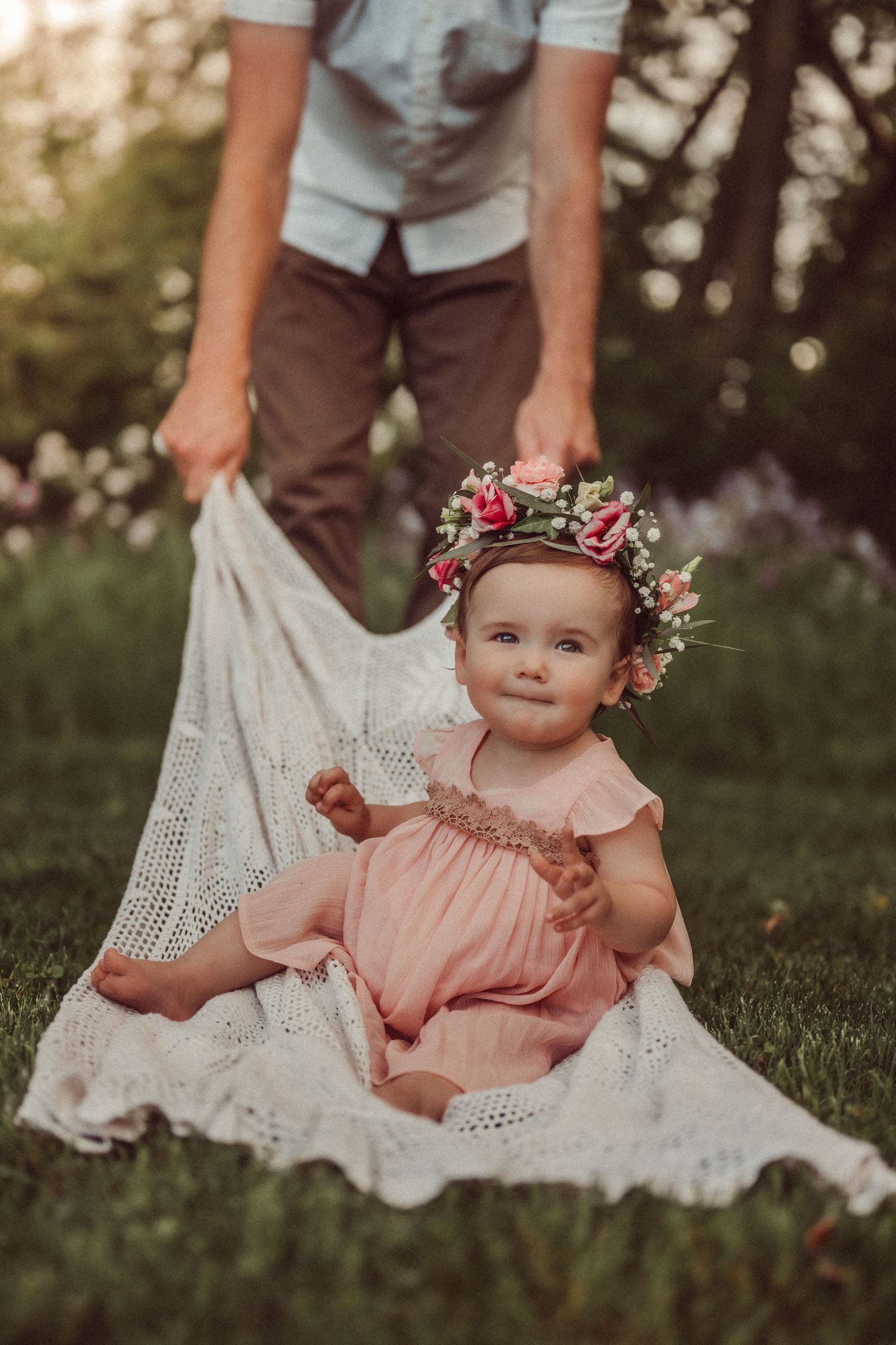 A small child in pink dress with floral crown sits on white lace blanket in garden setting.