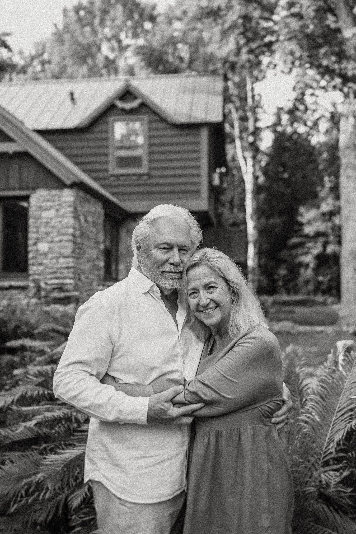 Black and white portrait of mature couple hugging and smiling outside their cabin home.