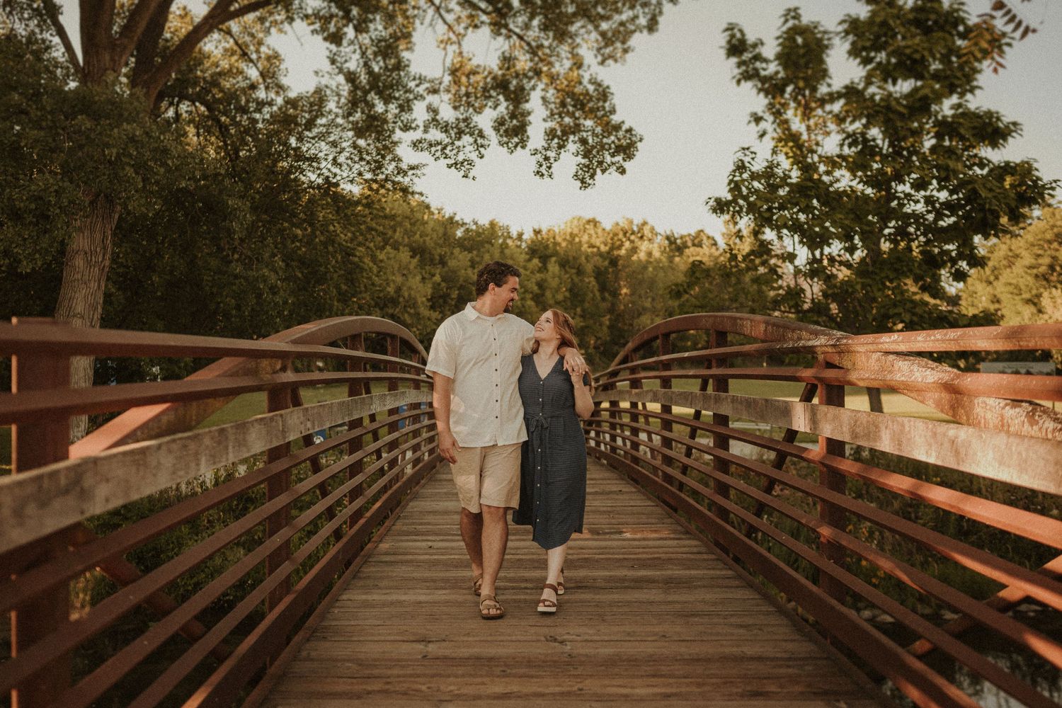 A romantic sunset photograph of a couple walking across a wooden bridge in a park.