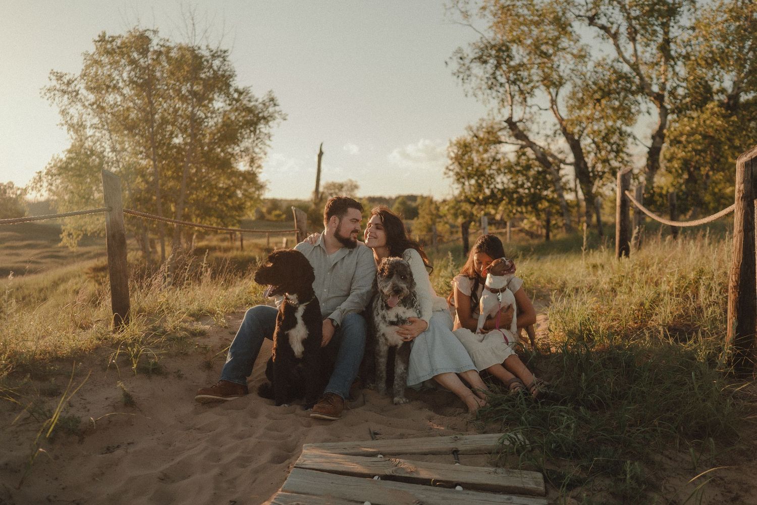 Family with dog sitting on wooden planks in golden sunlight, surrounded by trees and fields at sunset.
