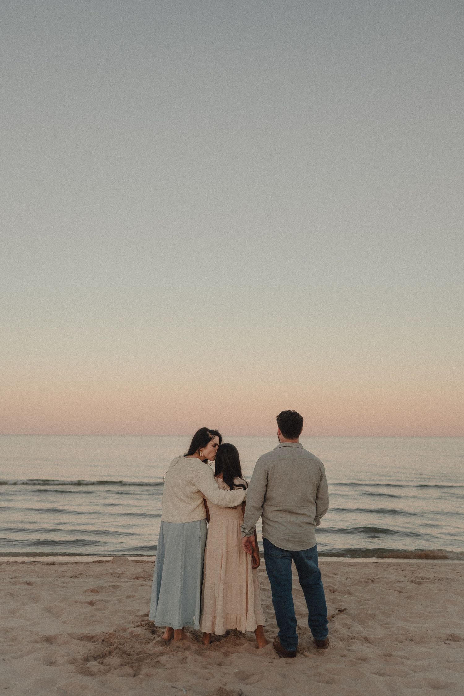Family of three standing together on a sandy beach at sunset, facing the calm ocean under a pastel sky.