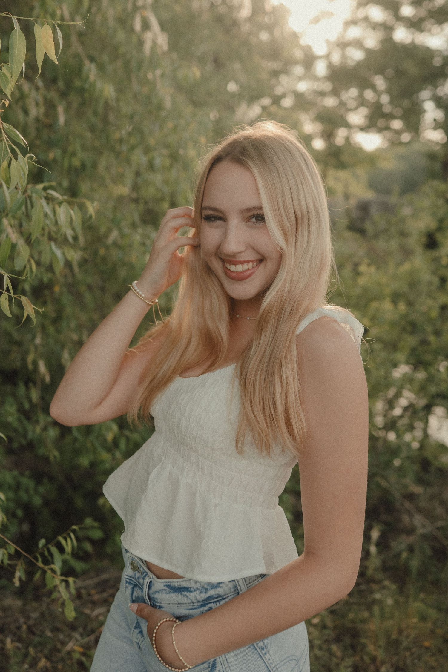 Smiling blonde woman in white crop top and jeans posing outdoors among green trees in golden hour light.