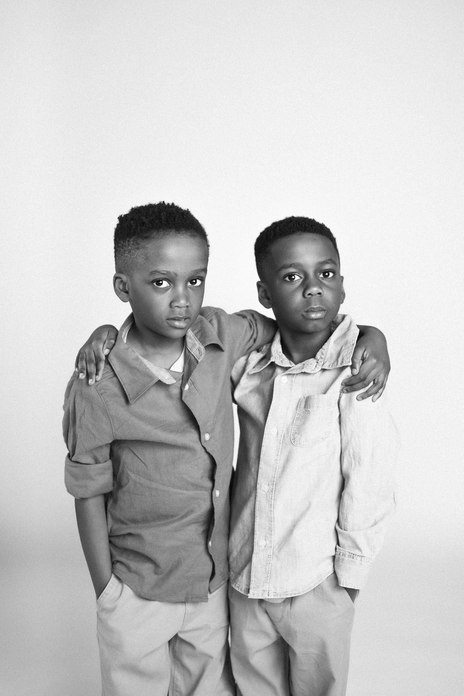 Two young boys in casual attire pose together against a white background in a black and white portrait series.