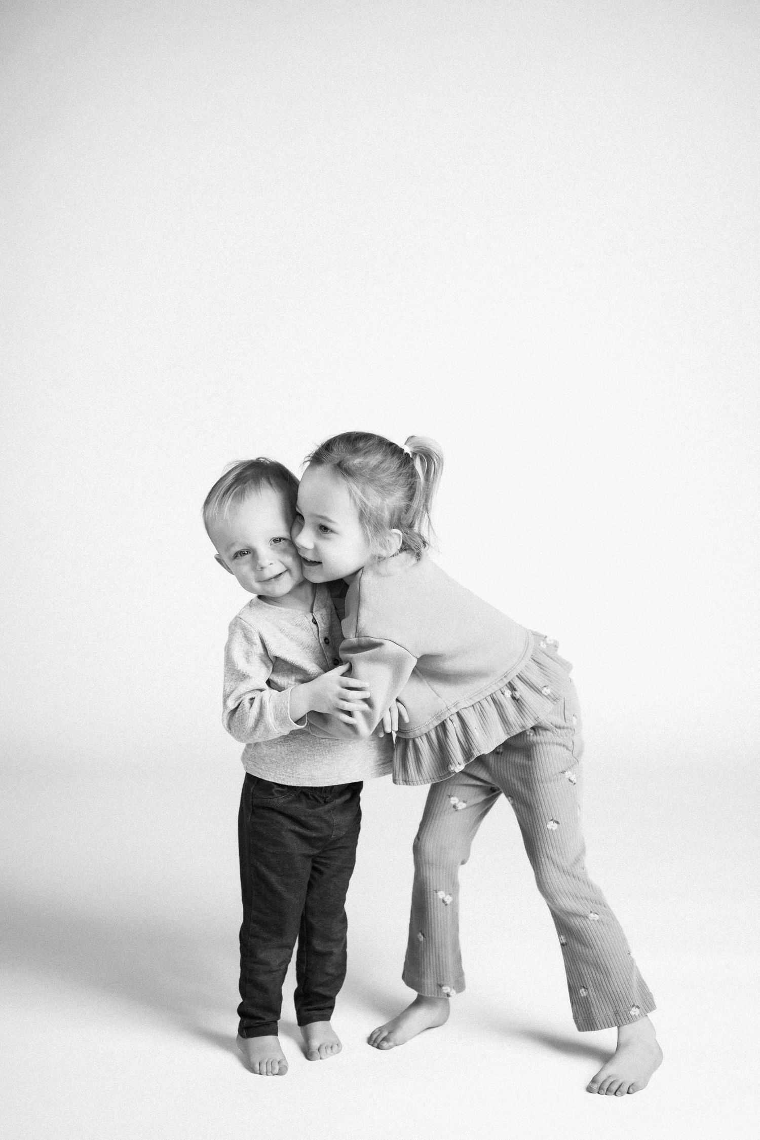 Two young children share a sweet candid moment in black and white against a plain background.