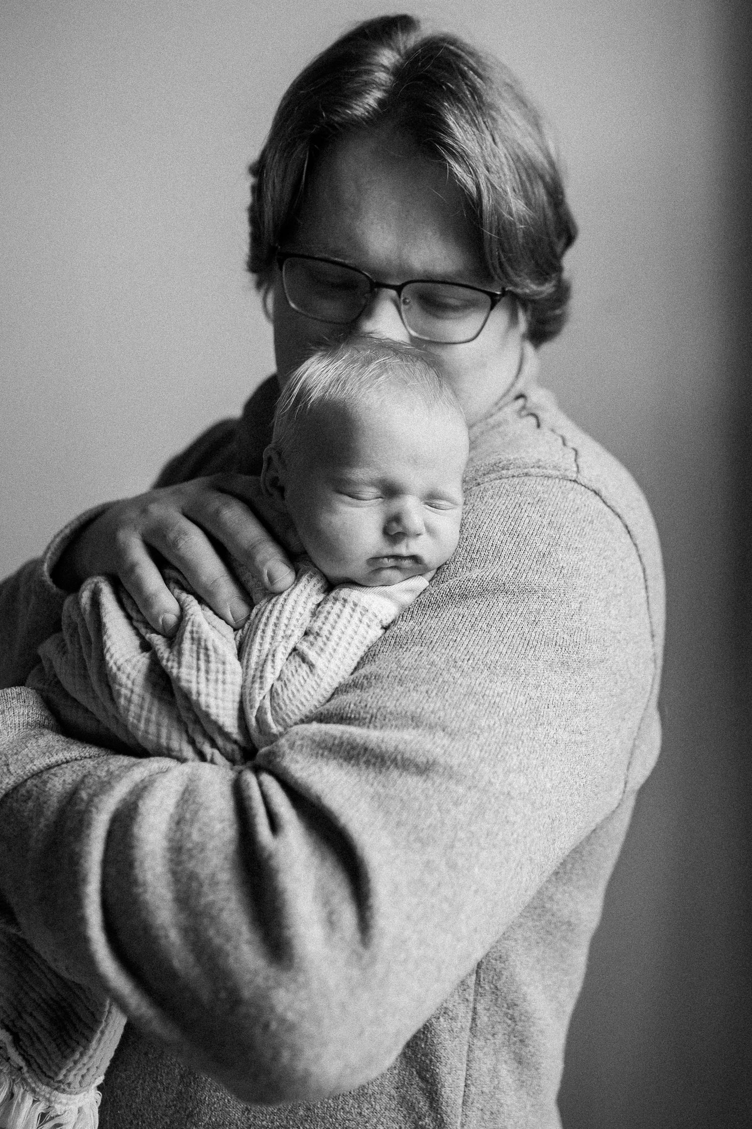 Black and white portrait of father in glasses gently cradling sleeping newborn against chest.