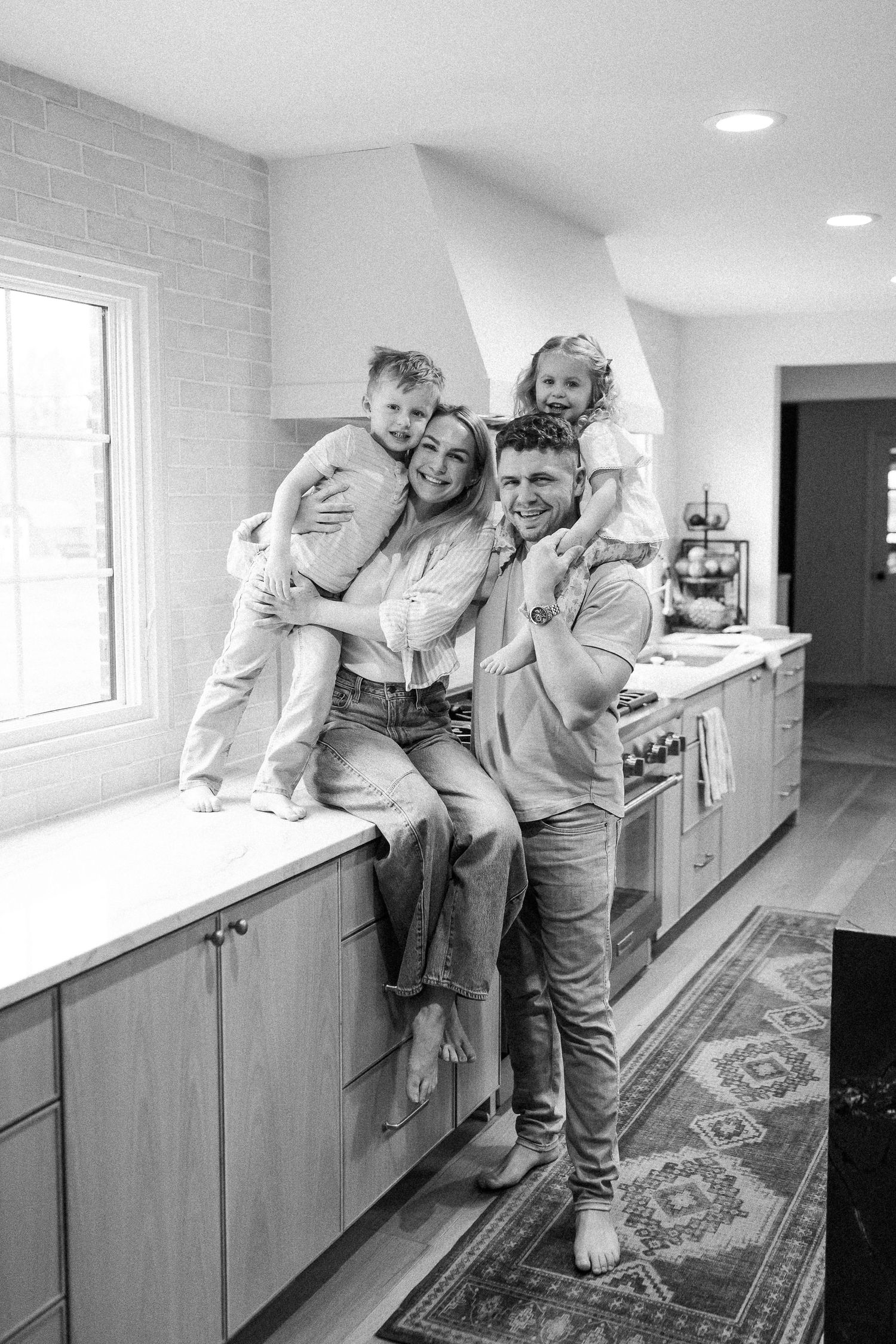 Black and white family portrait of parents and two young children posing happily in a modern kitchen.