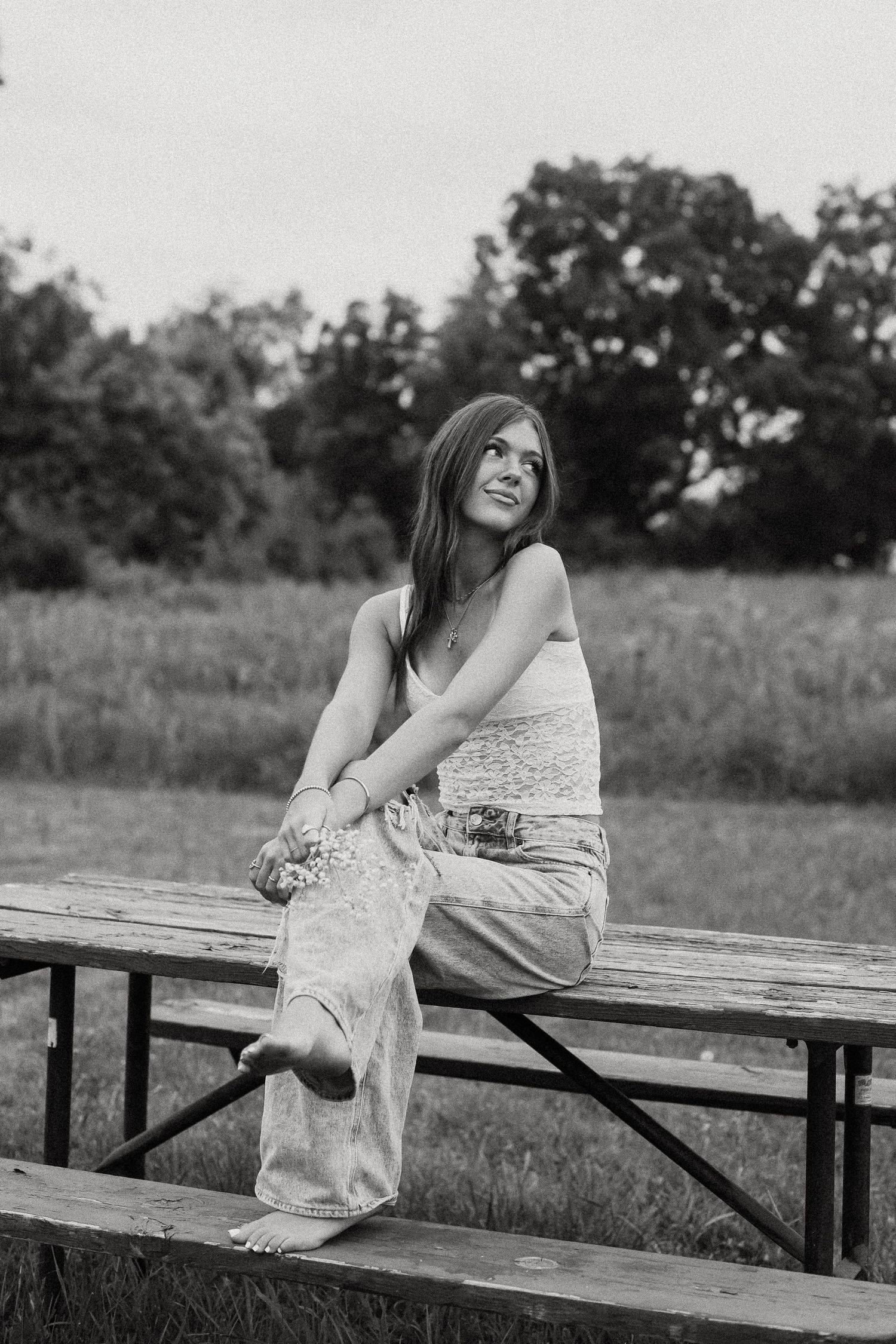Black and white photo of barefoot teenage girl sitting on wooden bleachers in open field.