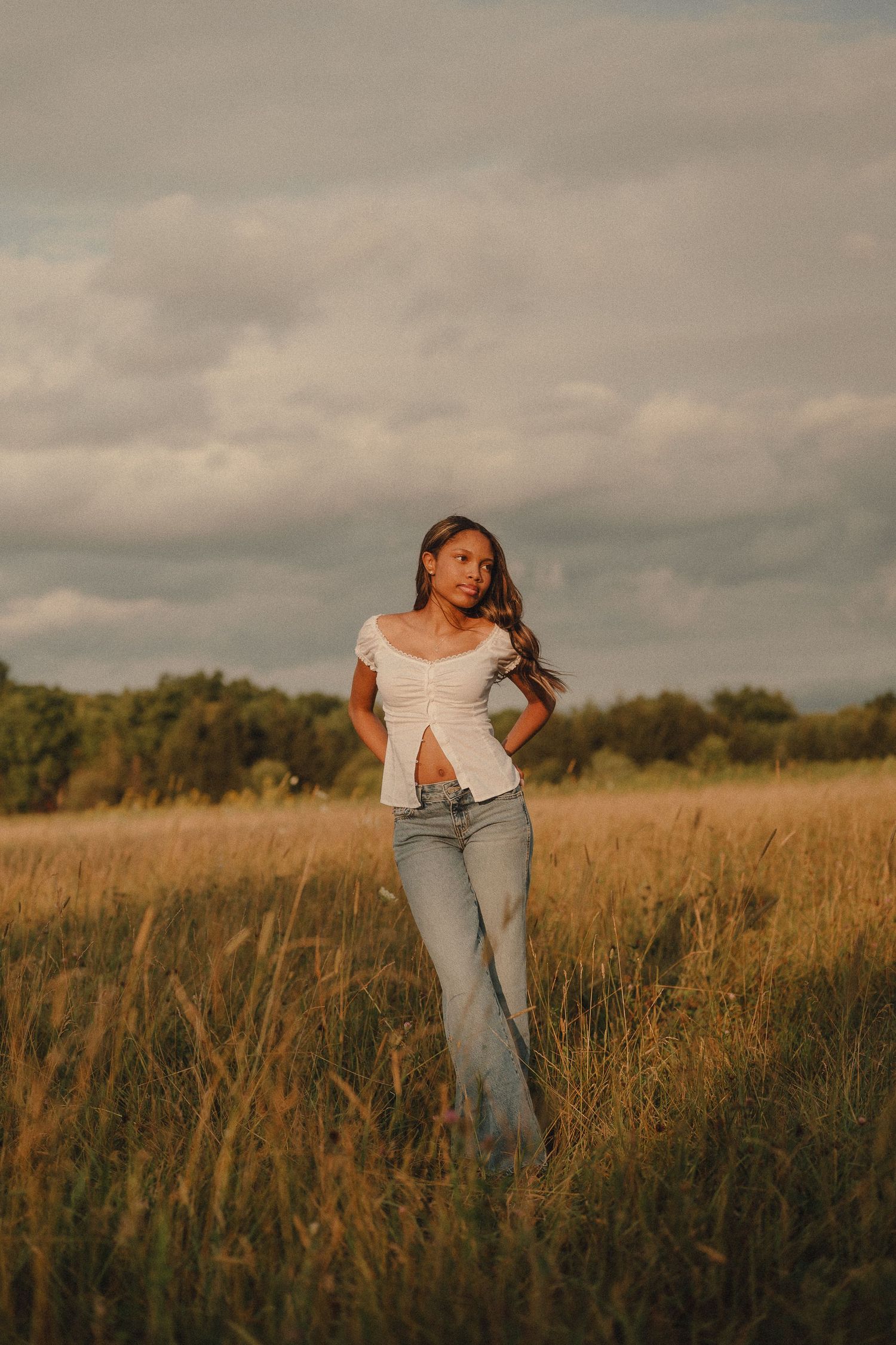 Young woman in white top and jeans poses in golden field at sunset.