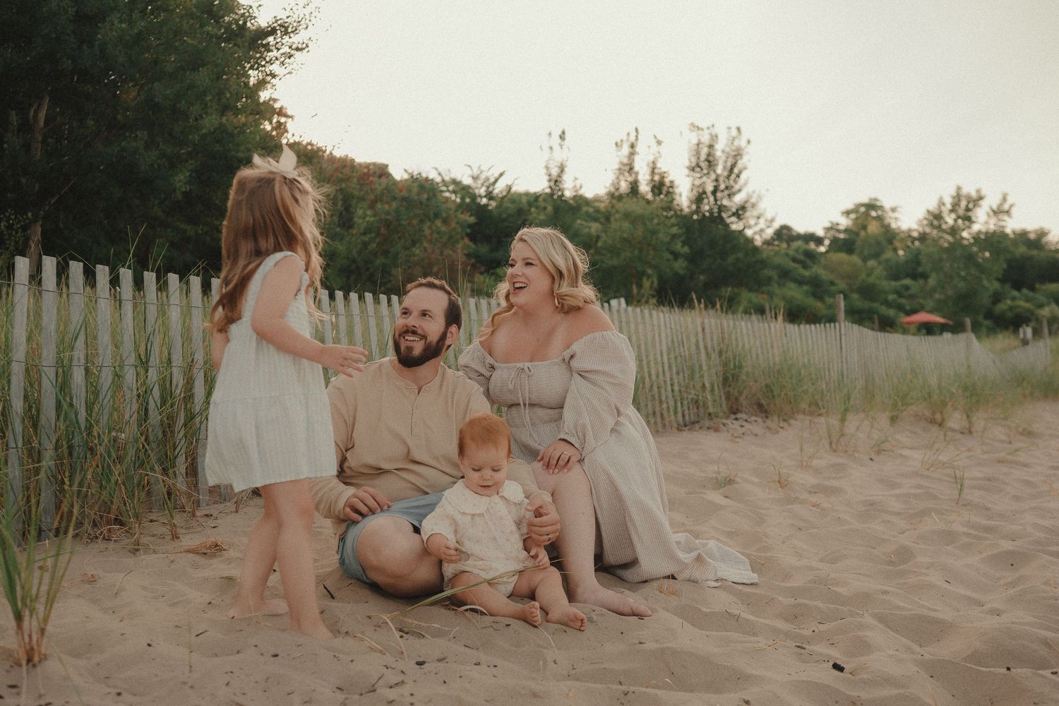 Family of four sitting on sandy beach near white fence, parents smiling at children in warm golden light.