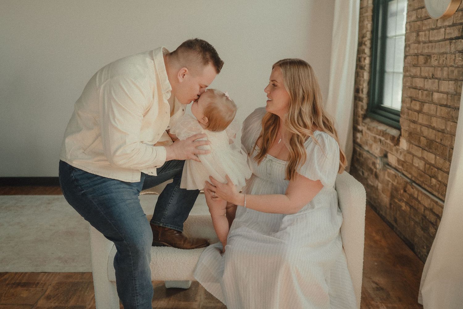 Parents lovingly kissing their baby girl in a bright studio with exposed brick wall.