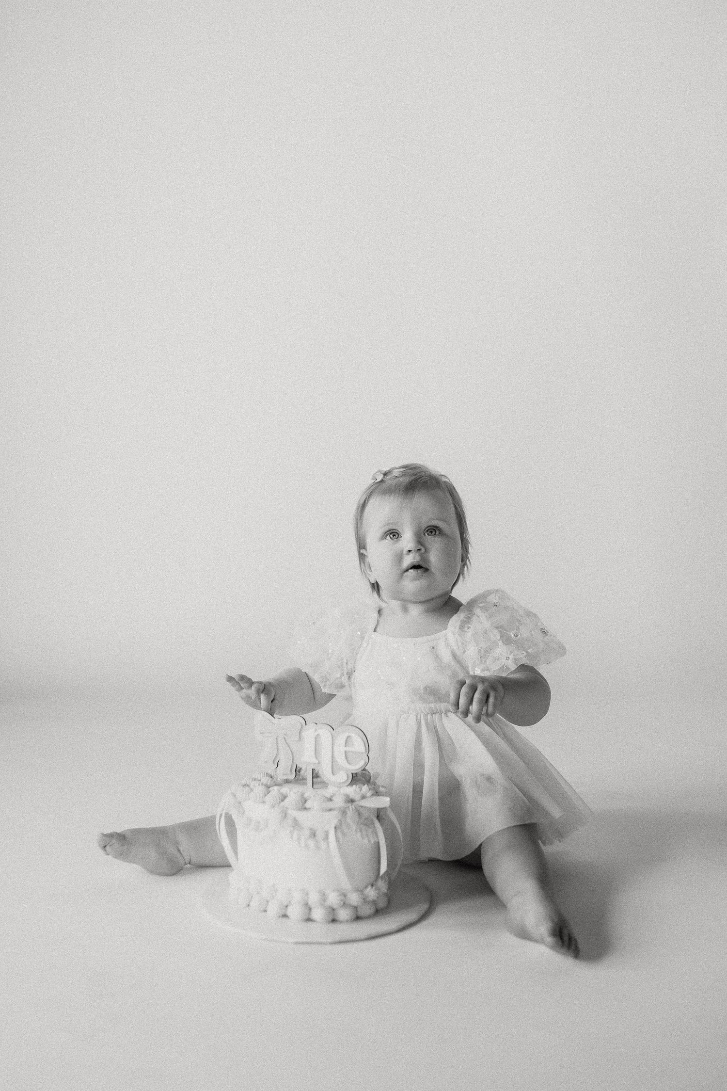Black and white photo of baby girl in lace dress beside first birthday cake with One topper in studio.