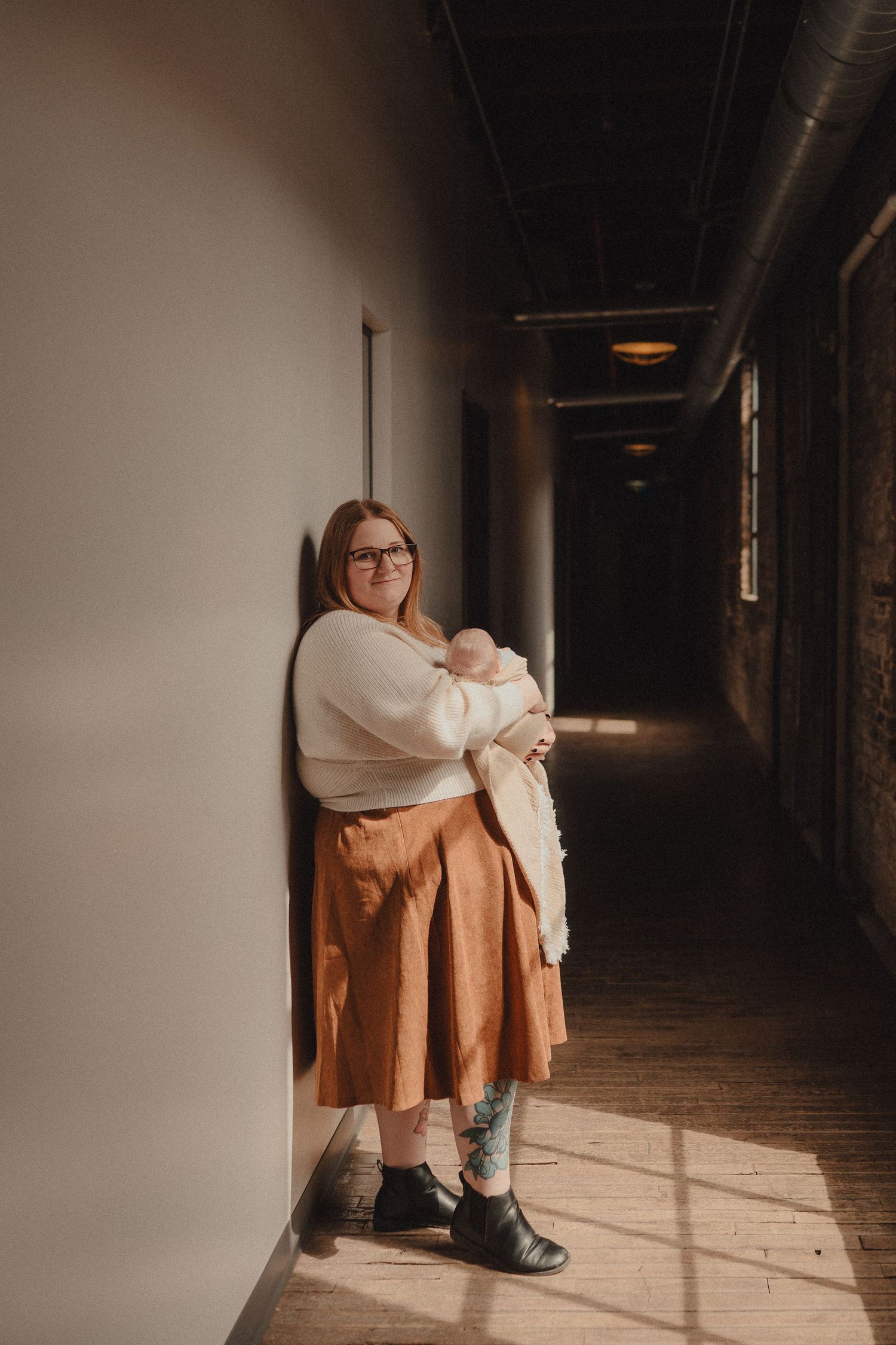 Mother in cream sweater and rust skirt leans against wall holding newborn in sunlit hallway.