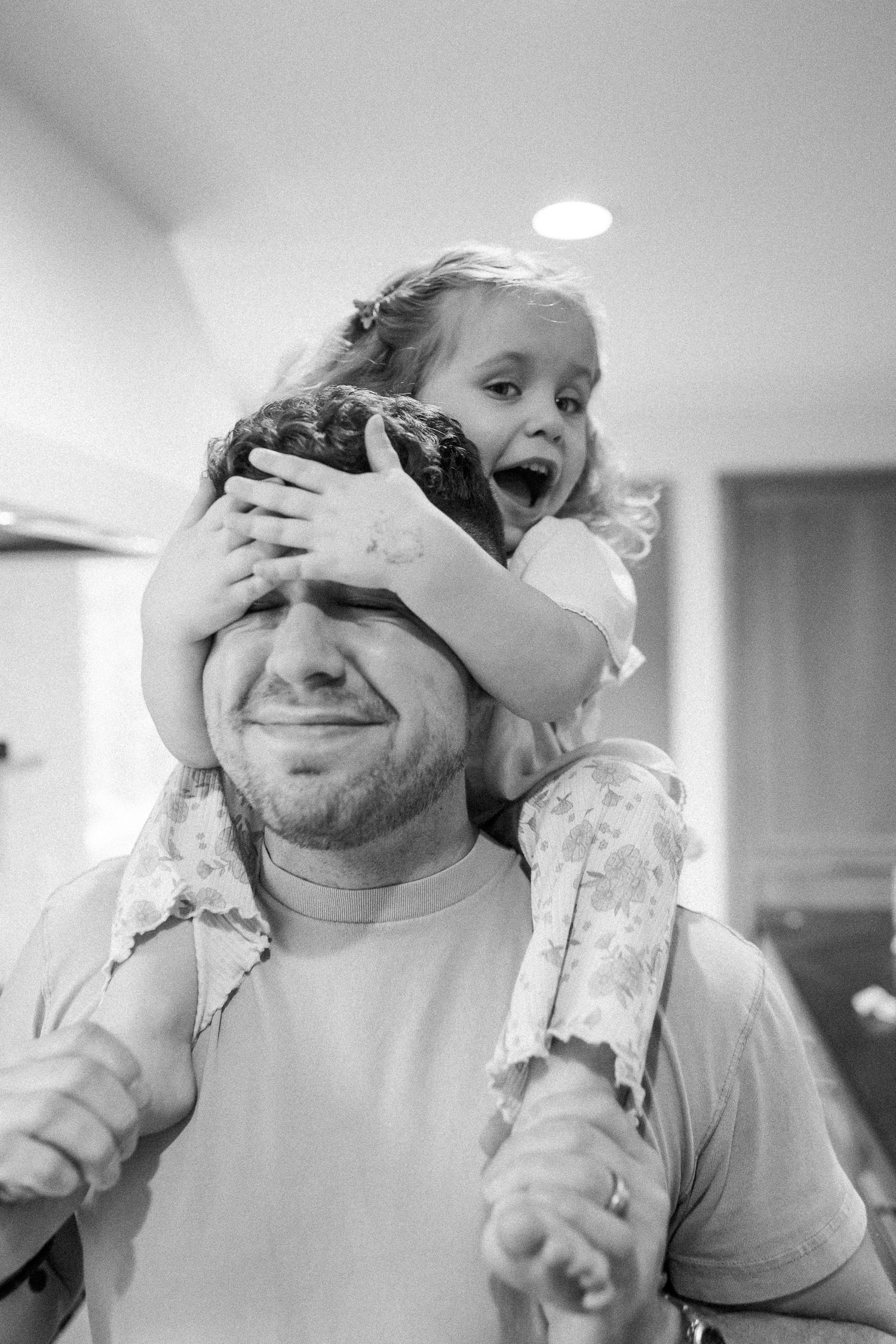 Black and white candid photo of a laughing girl covering her fathers eyes on his shoulders.