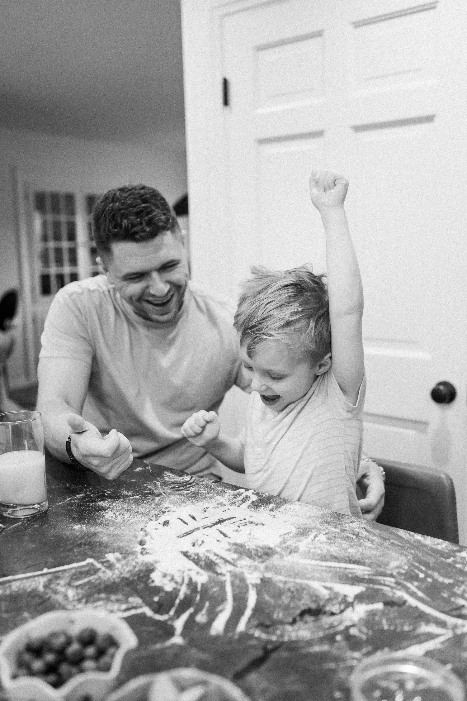 Black and white photo of a father laughing with his excited son while baking at a flour-covered table.