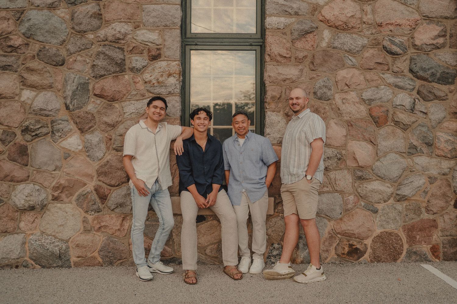 Four men smiling and posing together in front of a rustic stone wall near a window outdoors.