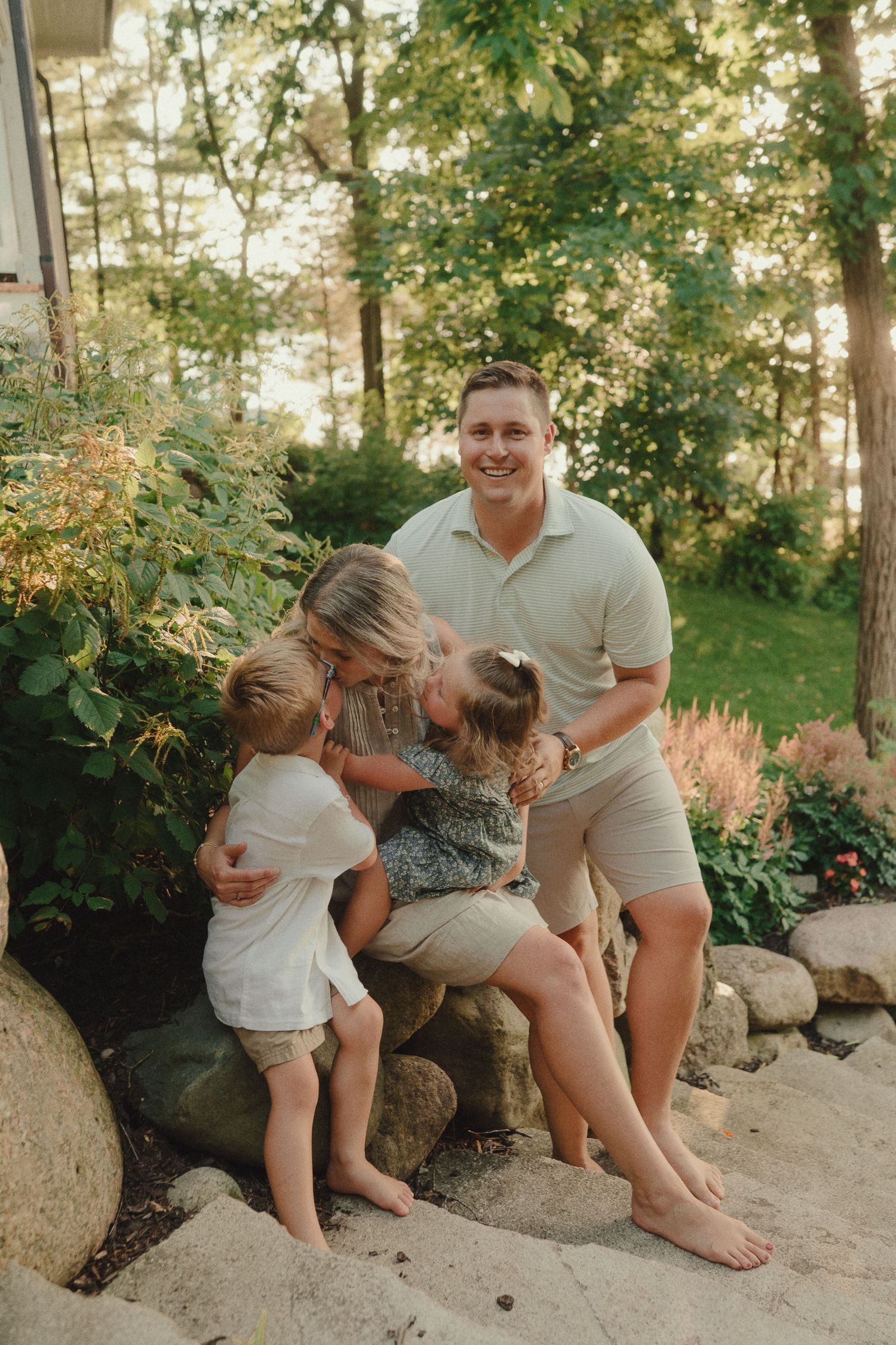 Happy family with two young children laughing together on stone steps surrounded by lush green garden.