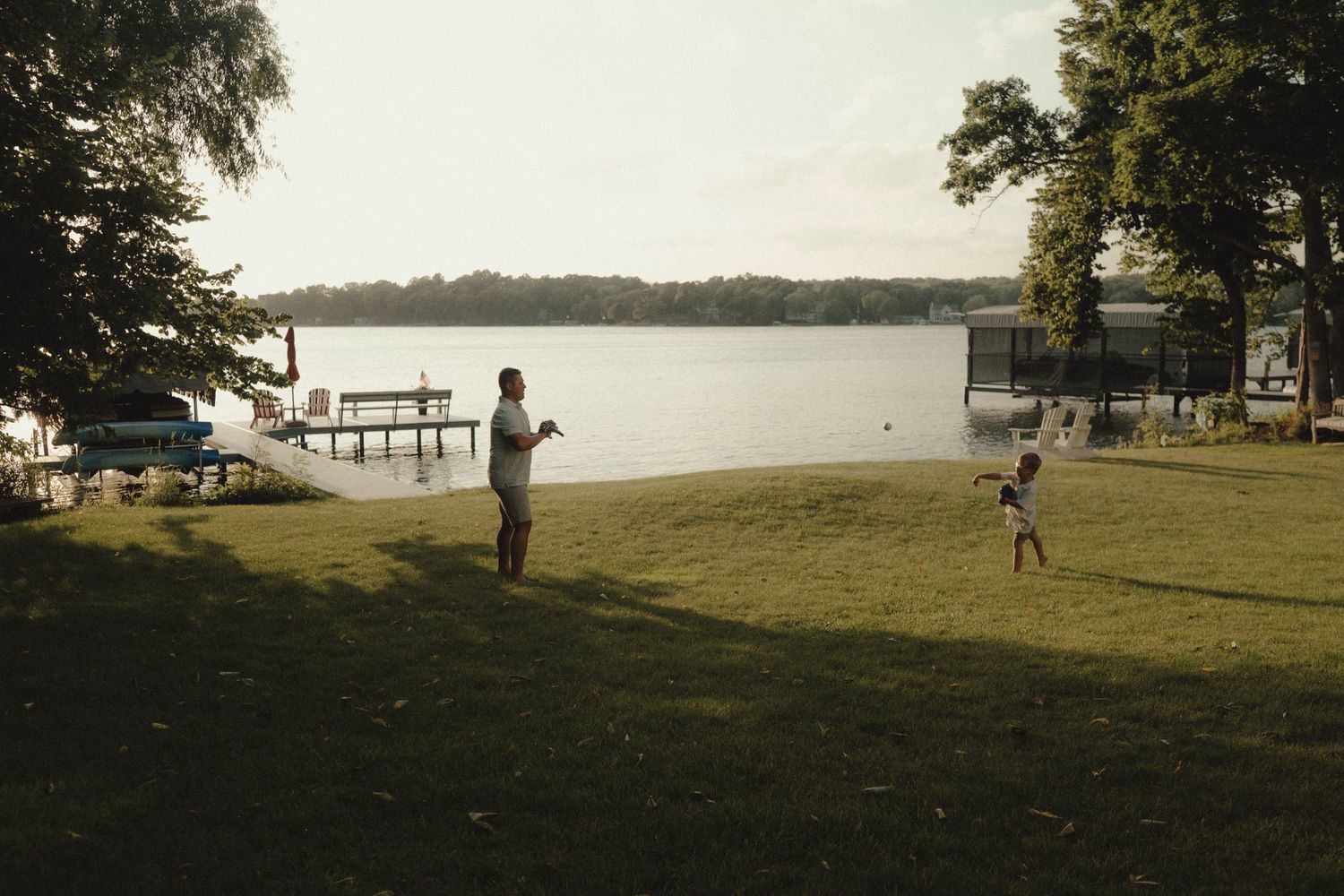 Two people play catch on a grassy lakeside yard at golden hour.
