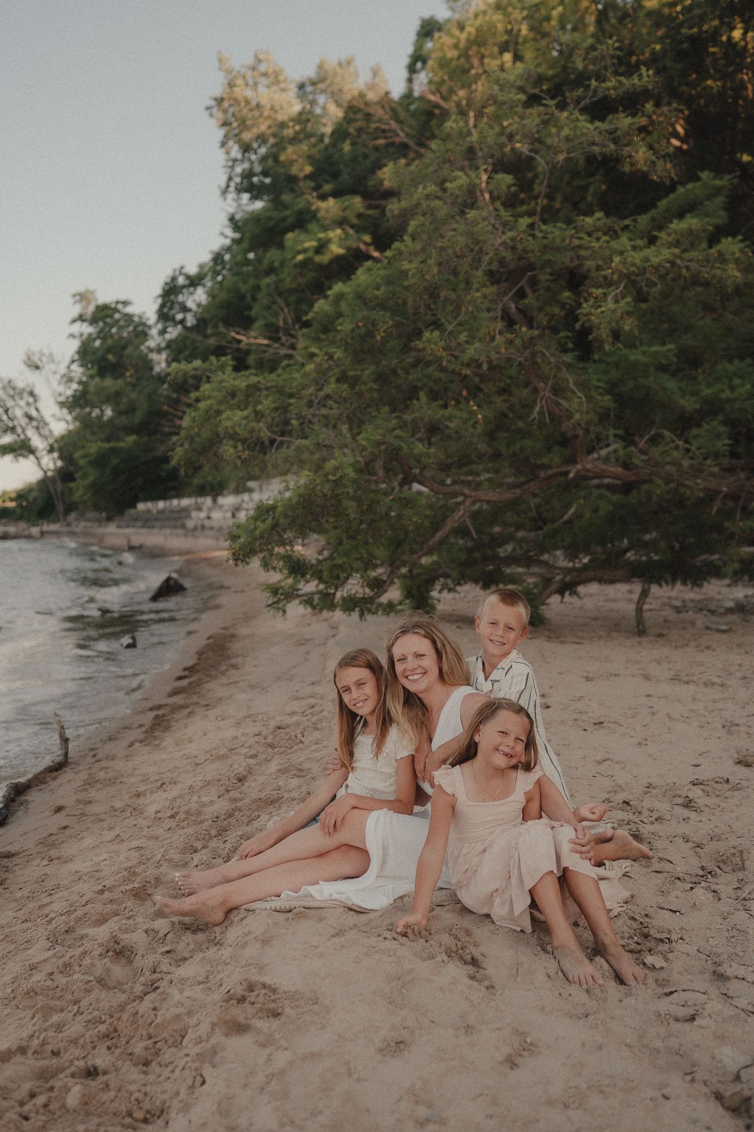 Mother and three children sit together on sandy beach with green trees behind them.