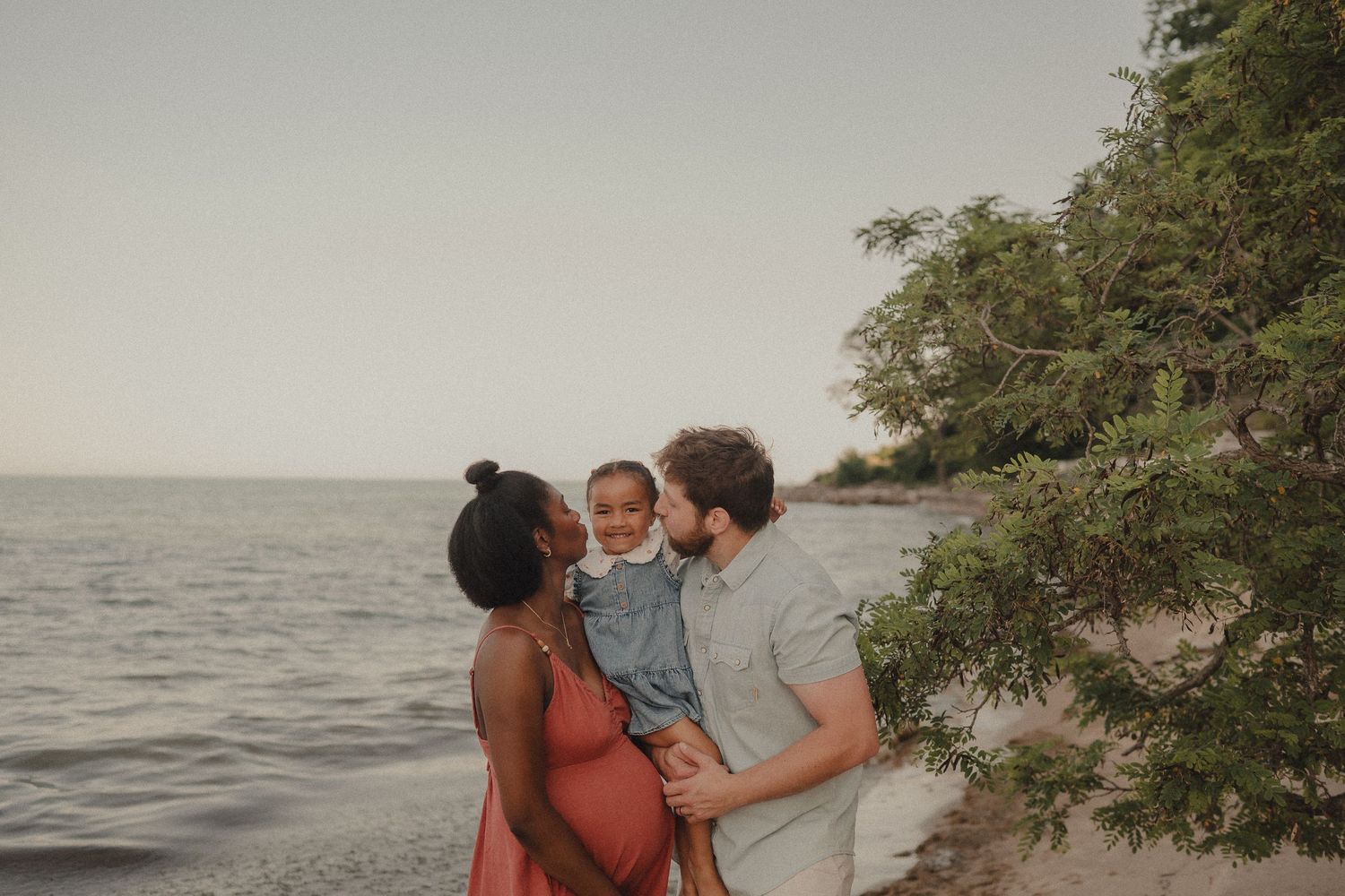 Pregnant woman and man kiss their toddler on a sandy beach near trees with calm water in the background.