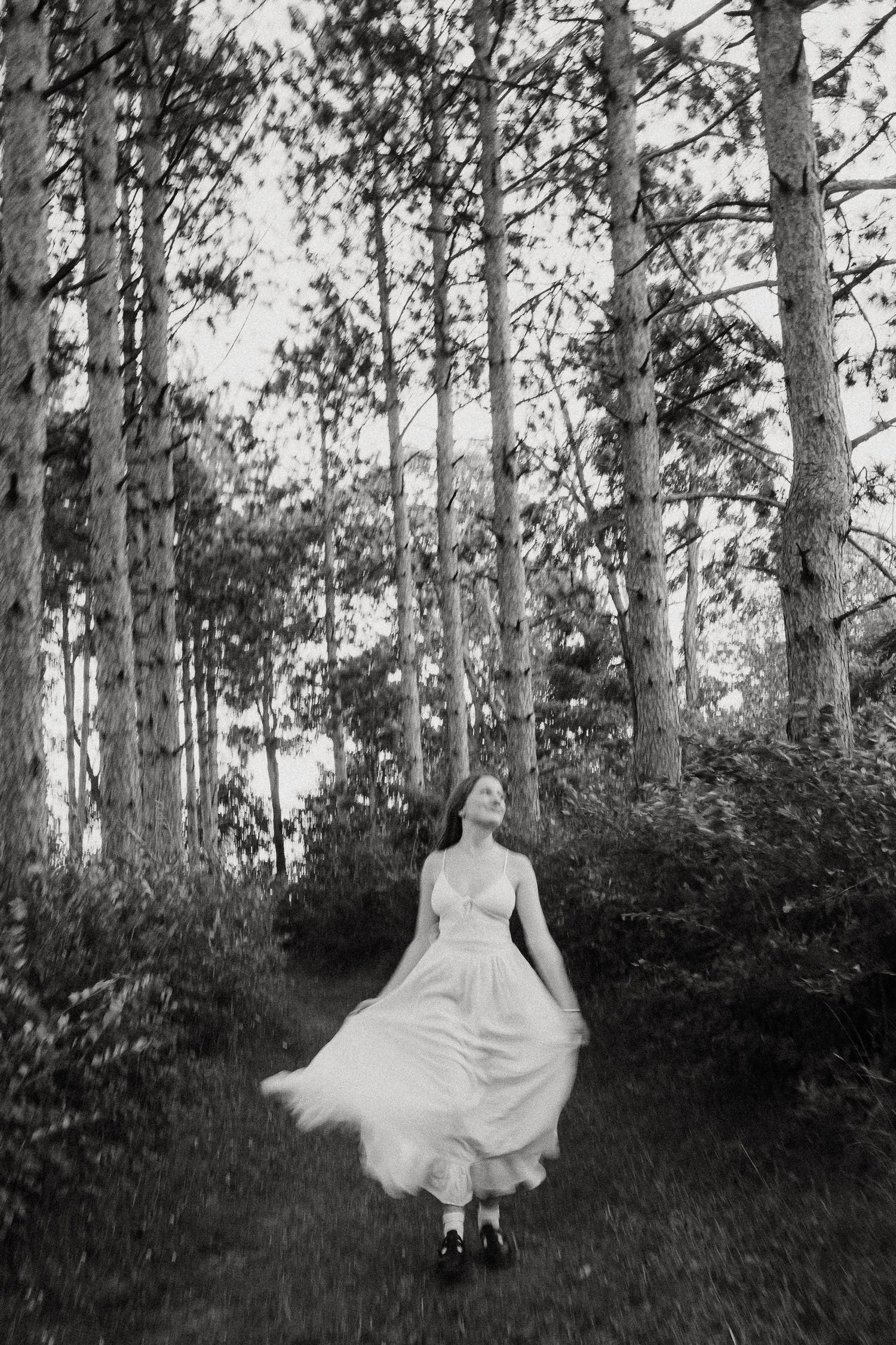 Black and white photo of woman twirling white dress in pine forest clearing.