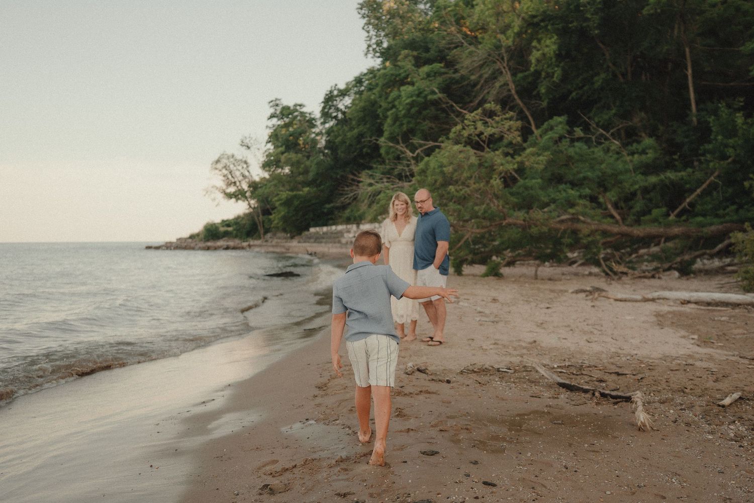Family walking barefoot on a sandy beach shore at sunset with lush green trees in the background.