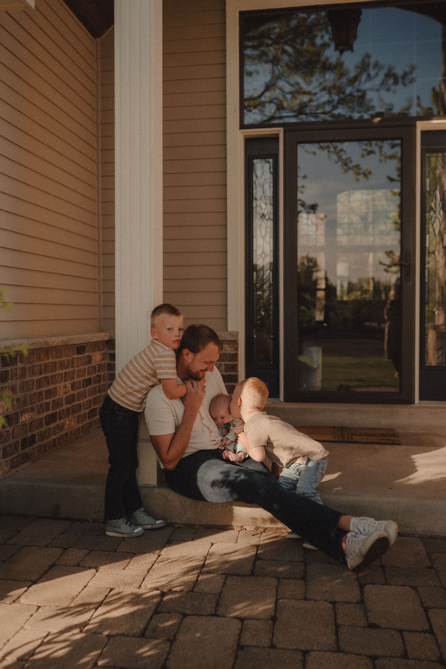 Father sitting on front porch steps with two young sons in warm golden sunset light.