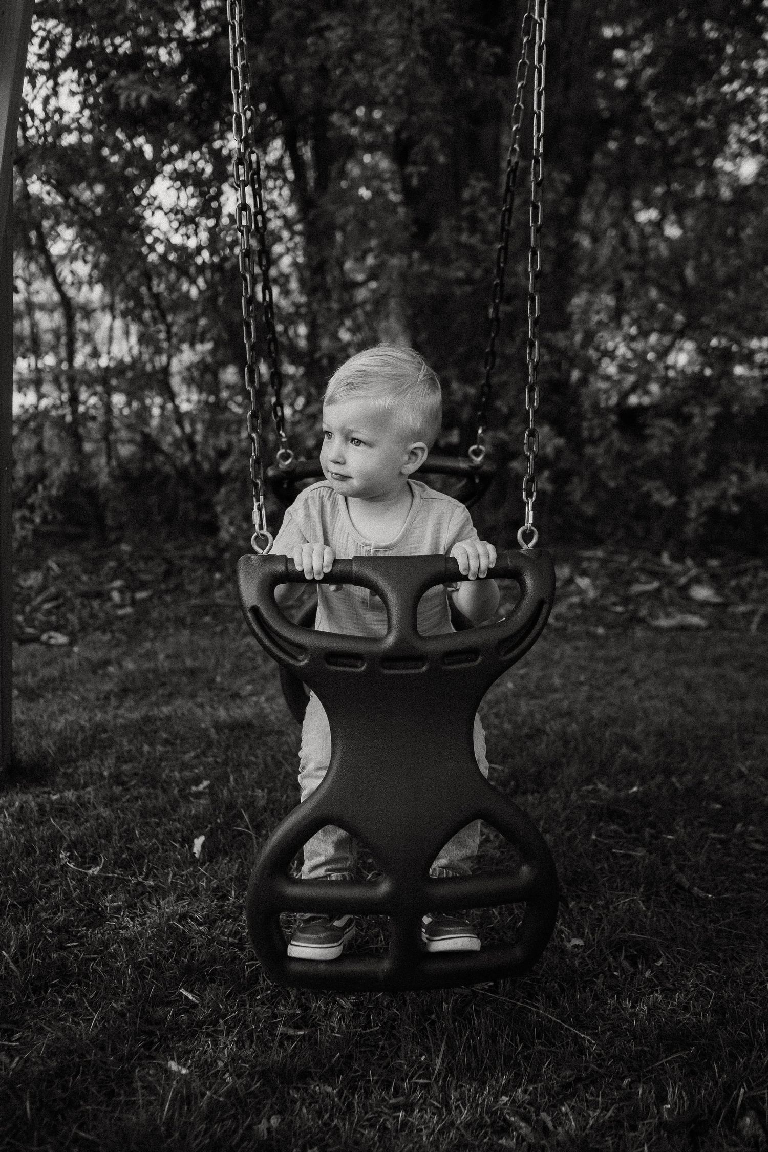 Cute blonde toddler sitting in outdoor bucket swing holding handlebar in black and white photo.