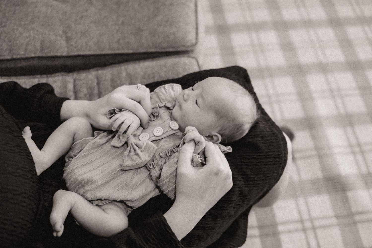 Black and white image of a baby lying on plaid bedding in soft window light.