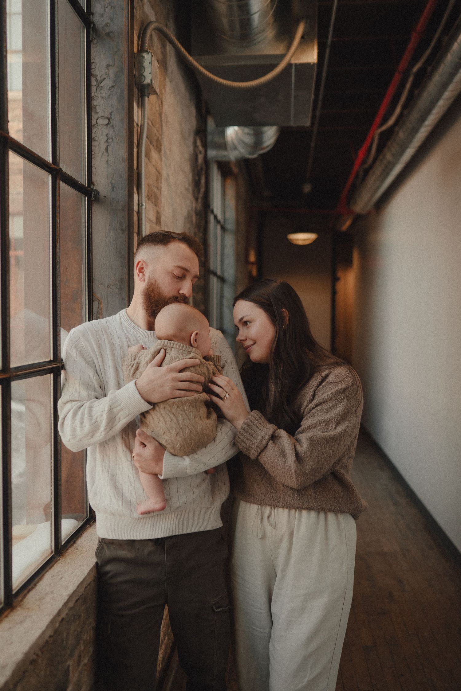 Family photo session in an industrial hallway with exposed brick and large windows creating dramatic lighting.
