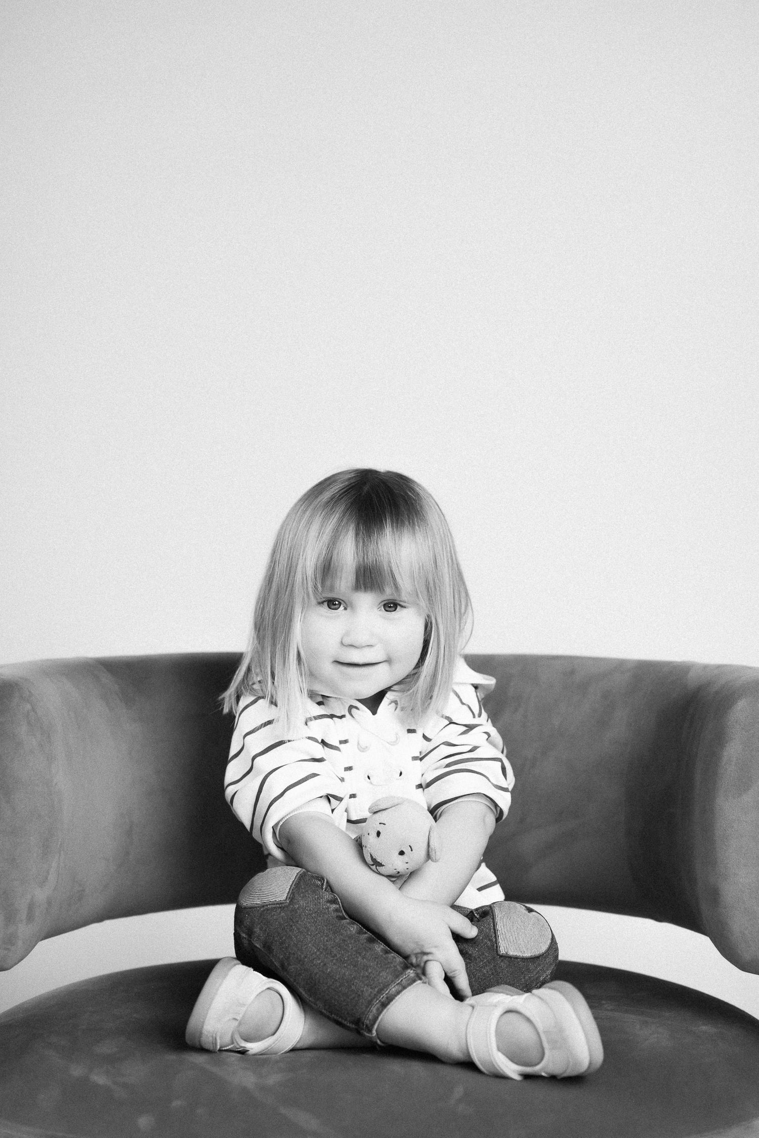 A young child in striped clothing sits cross-legged on a gray couch in a black and white portrait.