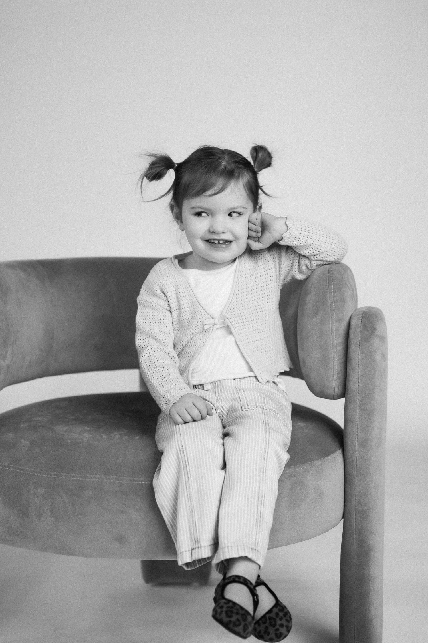 Child in white sweater and striped pants sitting casually on gray couch in black and white photo series.