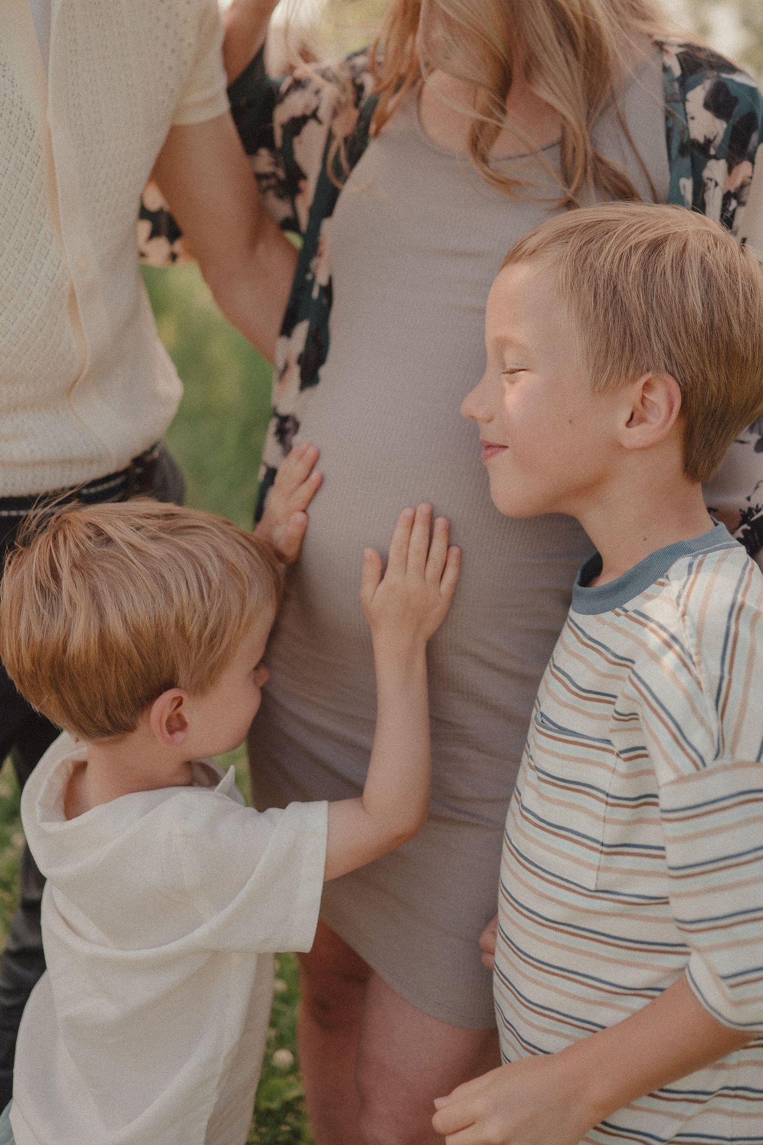 Two young brothers gently touching their pregnant mothers baby bump outdoors in warm natural light.