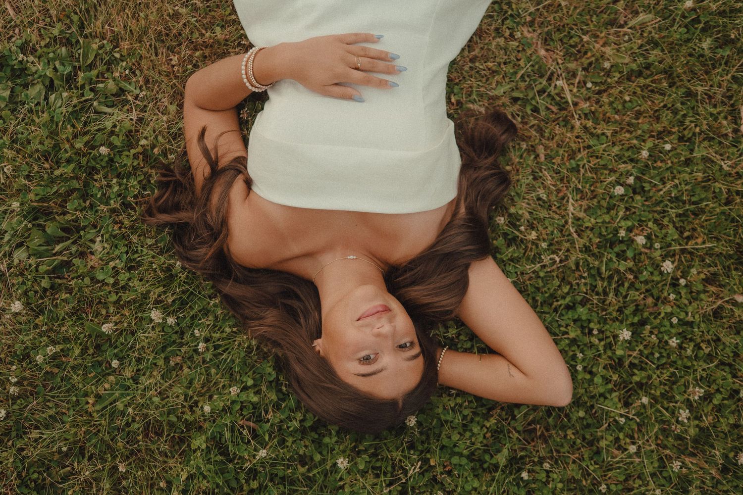 Young woman with long brown hair lying on green grass wearing a white dress, viewed from above.