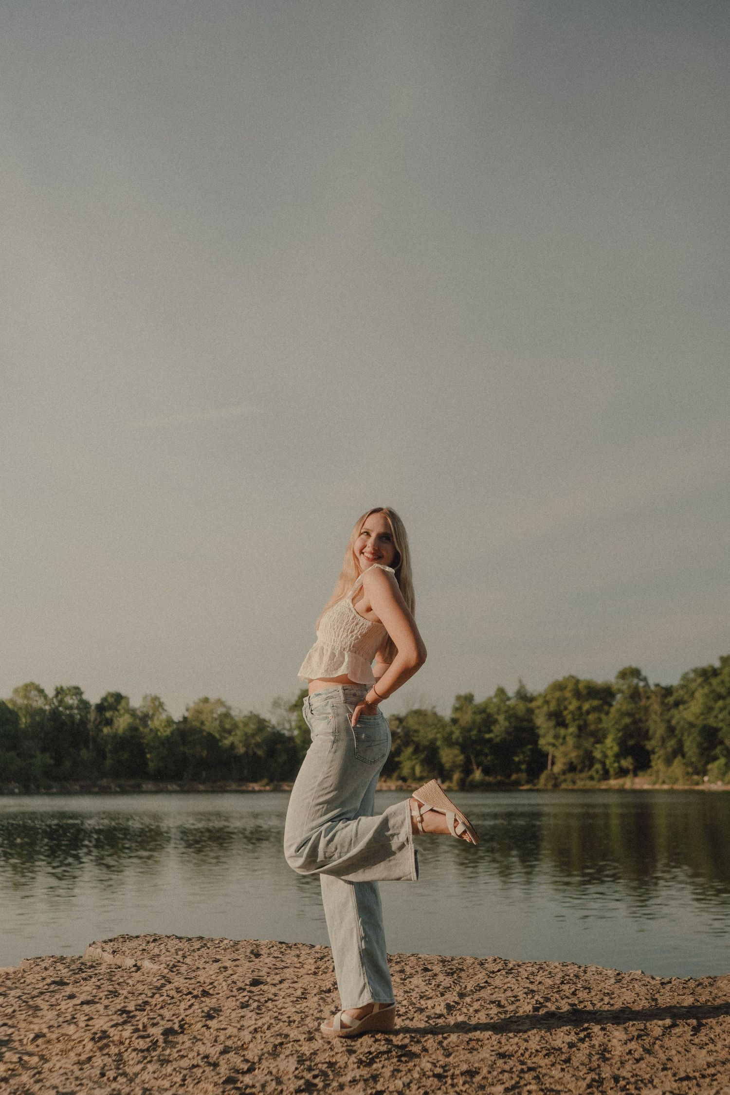 Young woman in light jeans and tank top posing by a serene lake with lush green trees in the background.
