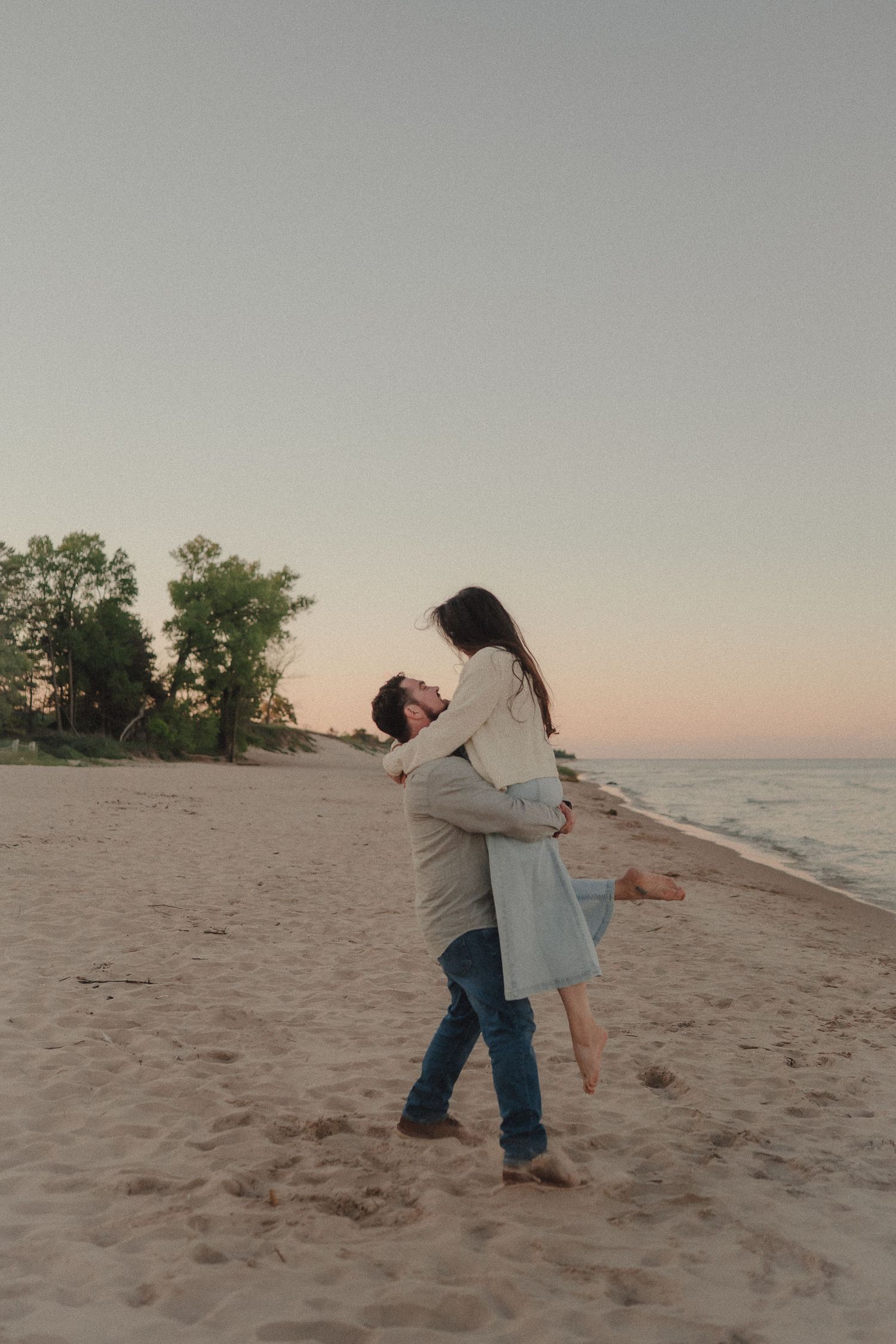 Man lifts woman in blue dress on sandy beach at dusk with trees behind them.