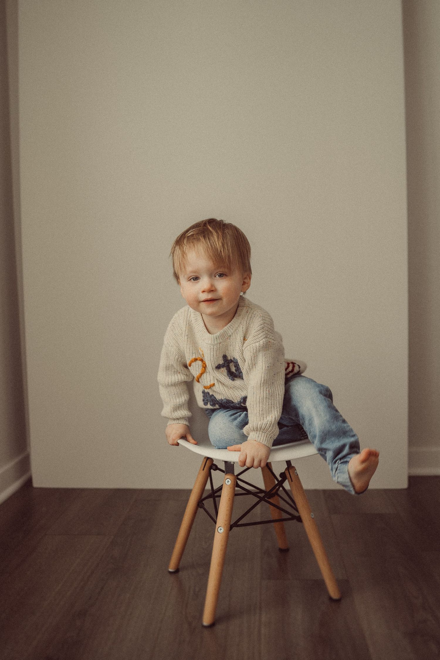 Child perched playfully on modern wooden chair against neutral wall background.