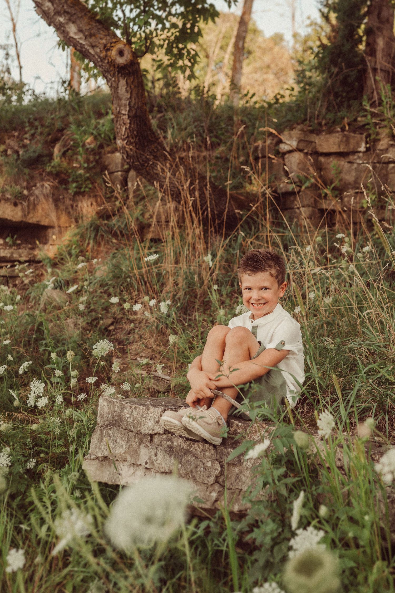 Child sits relaxed on stone ledge surrounded by wildflowers in warm evening light.