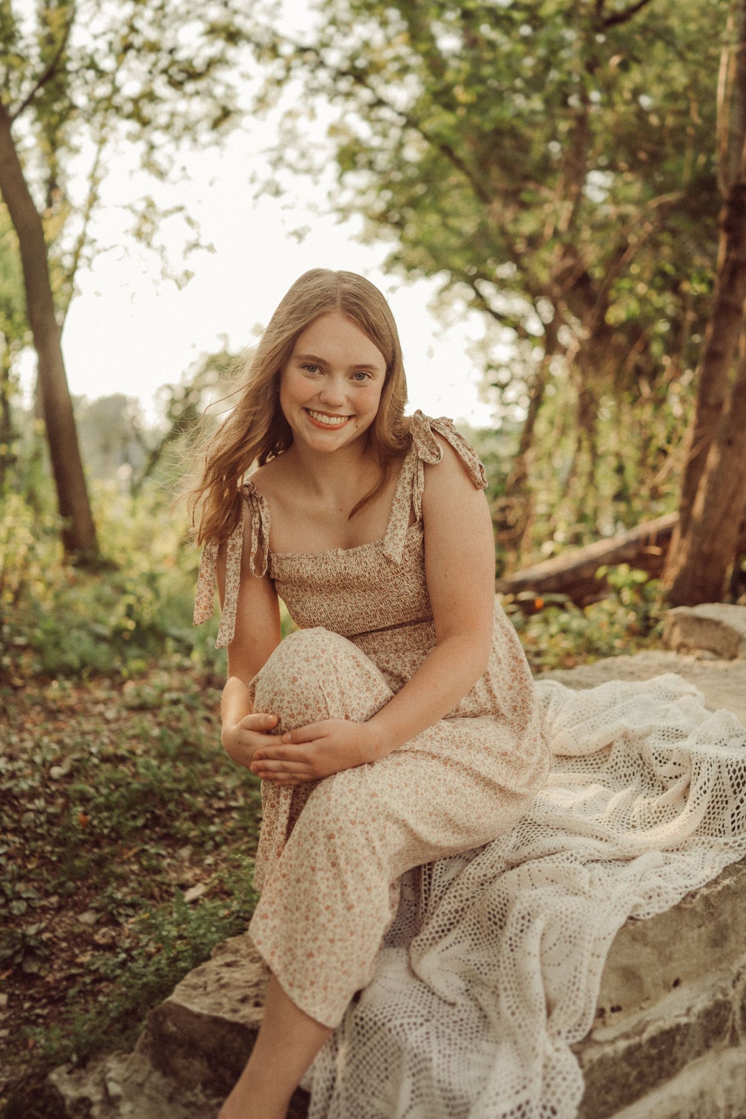 Woman in vintage-inspired dress sits on stone steps in a wooded setting during golden hour.