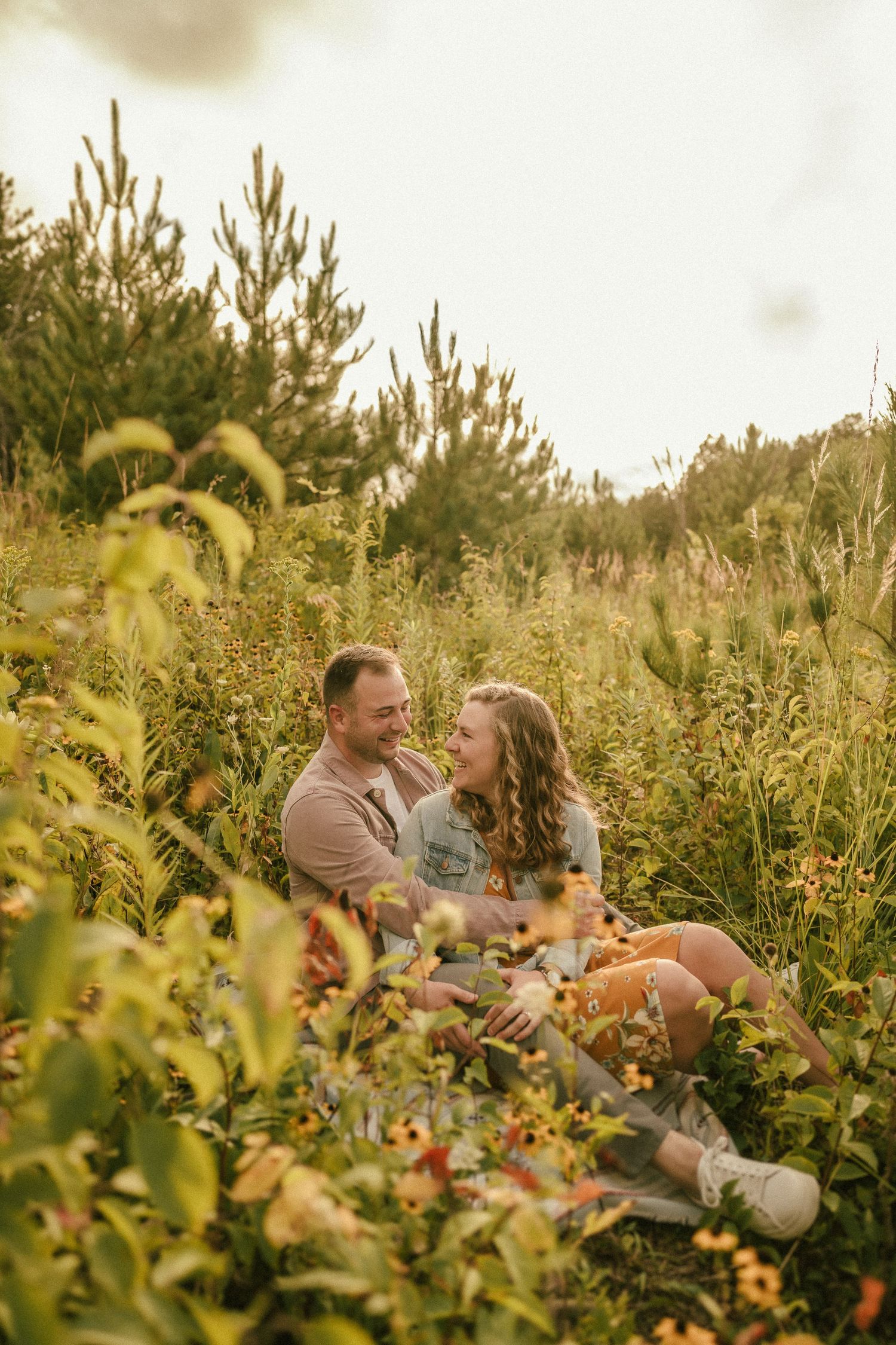 A couple sits together in a field of wildflowers during golden hour.