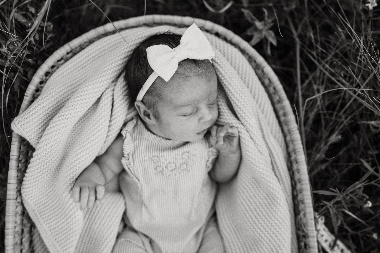 Black and white portrait of sleeping infant in white outfit lying in woven bassinet.