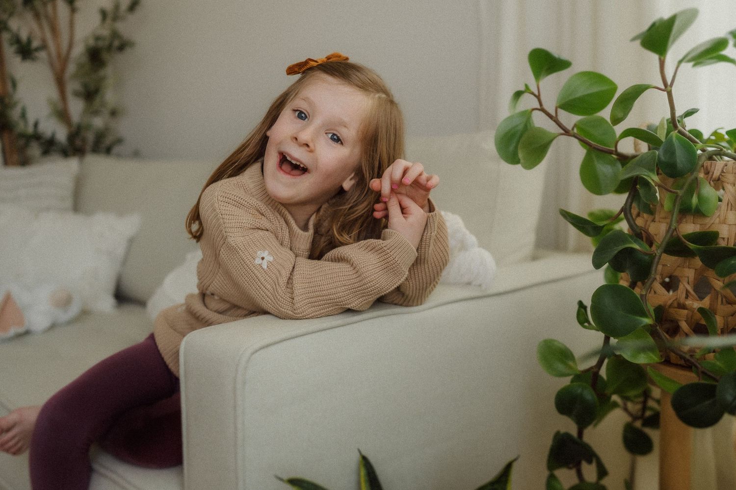 Playful child in beige sweater and purple leggings lounging on white couch next to houseplants.