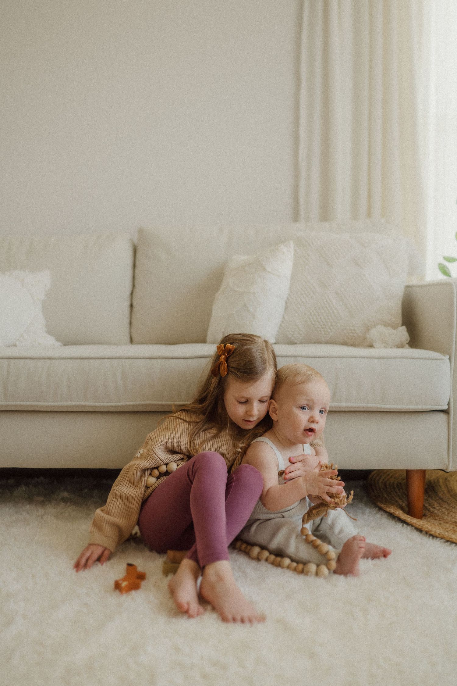 Two siblings share a sweet moment while sitting on a plush carpet near a beige couch.