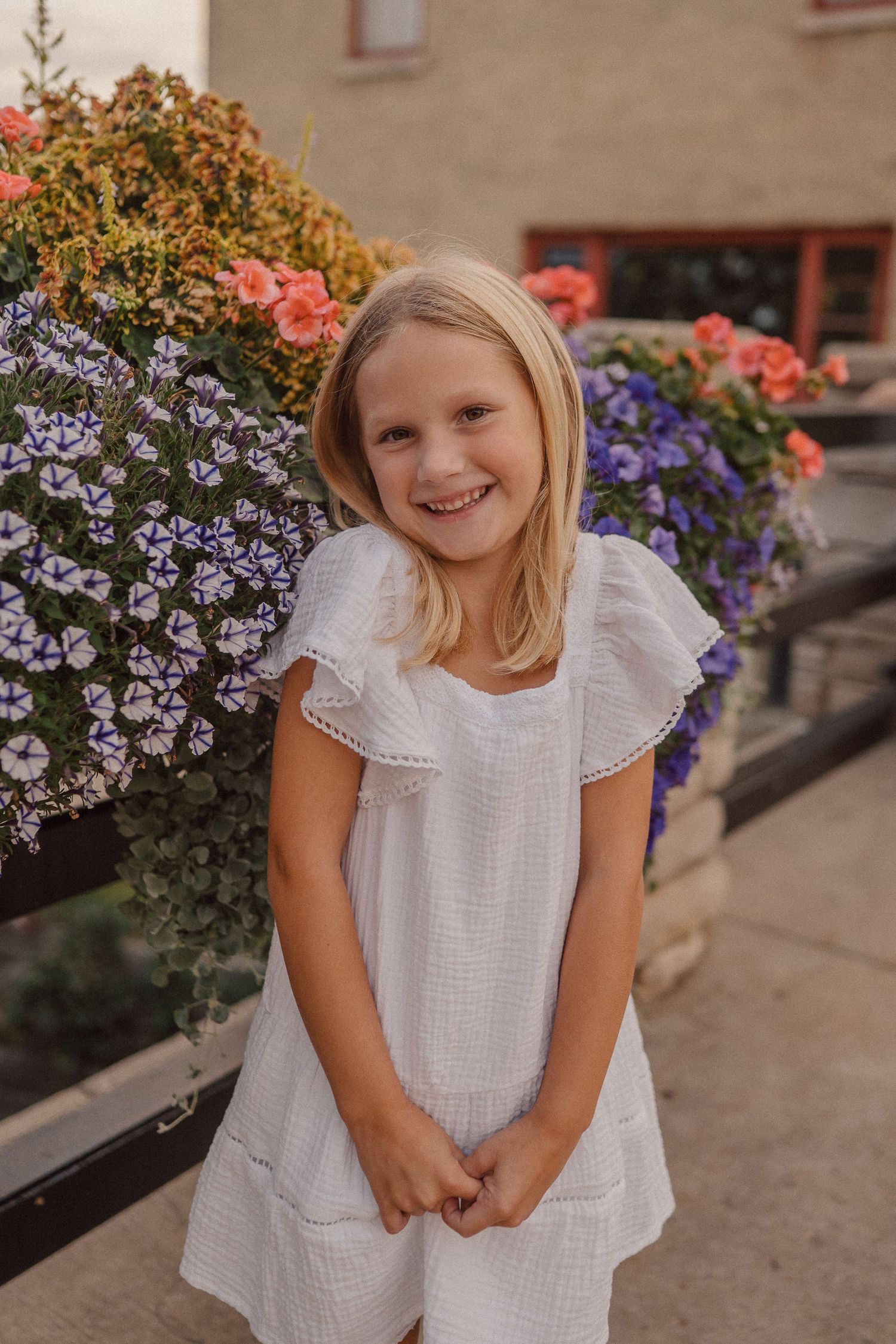 Young girl in white dress smiles playfully near colorful garden flowers.