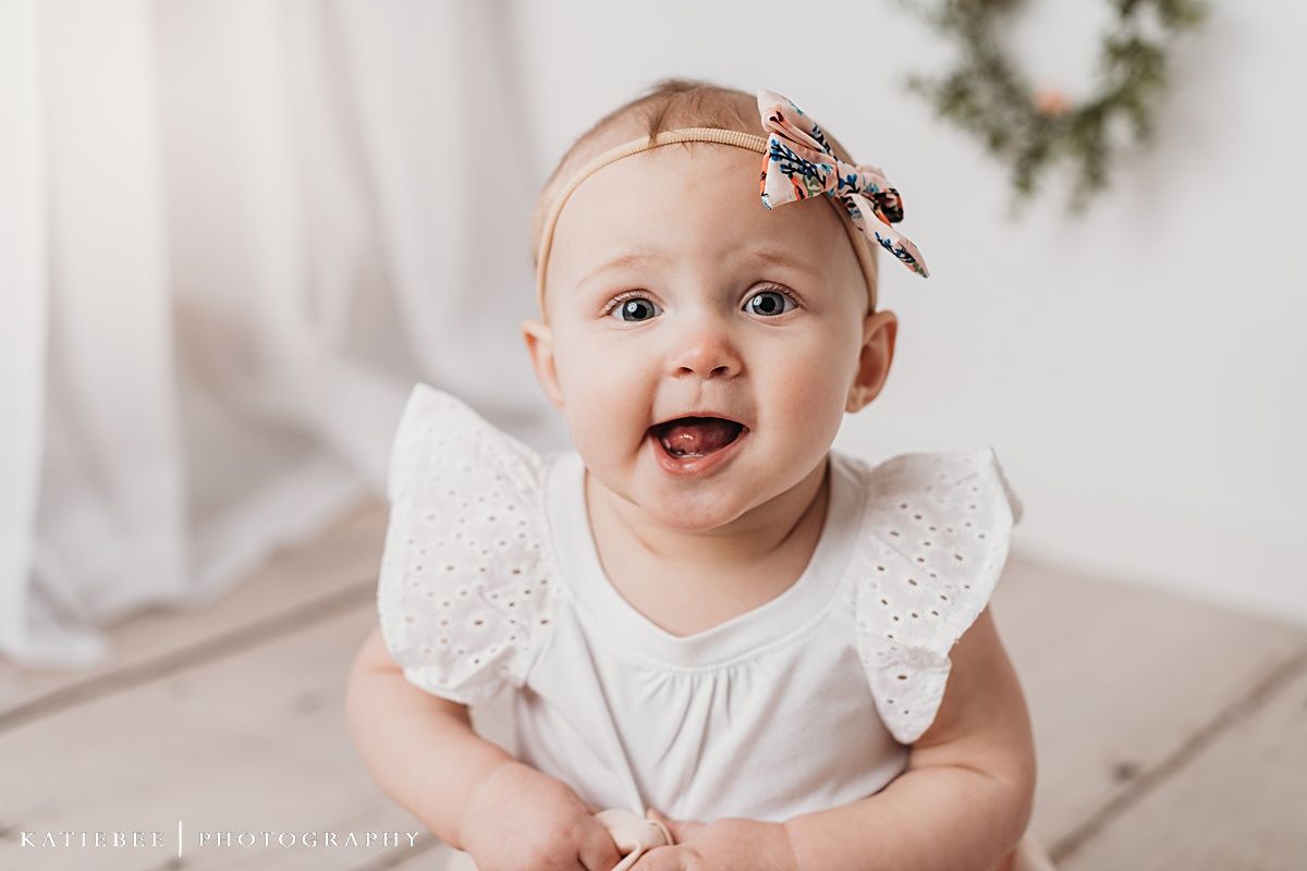 Baby wearing white eyelet dress and bow smiling at camera against white backdrop with wreaths.