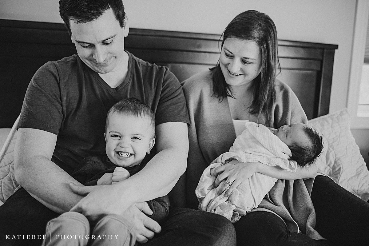 Black and white family portrait on bed with toddler and newborn.