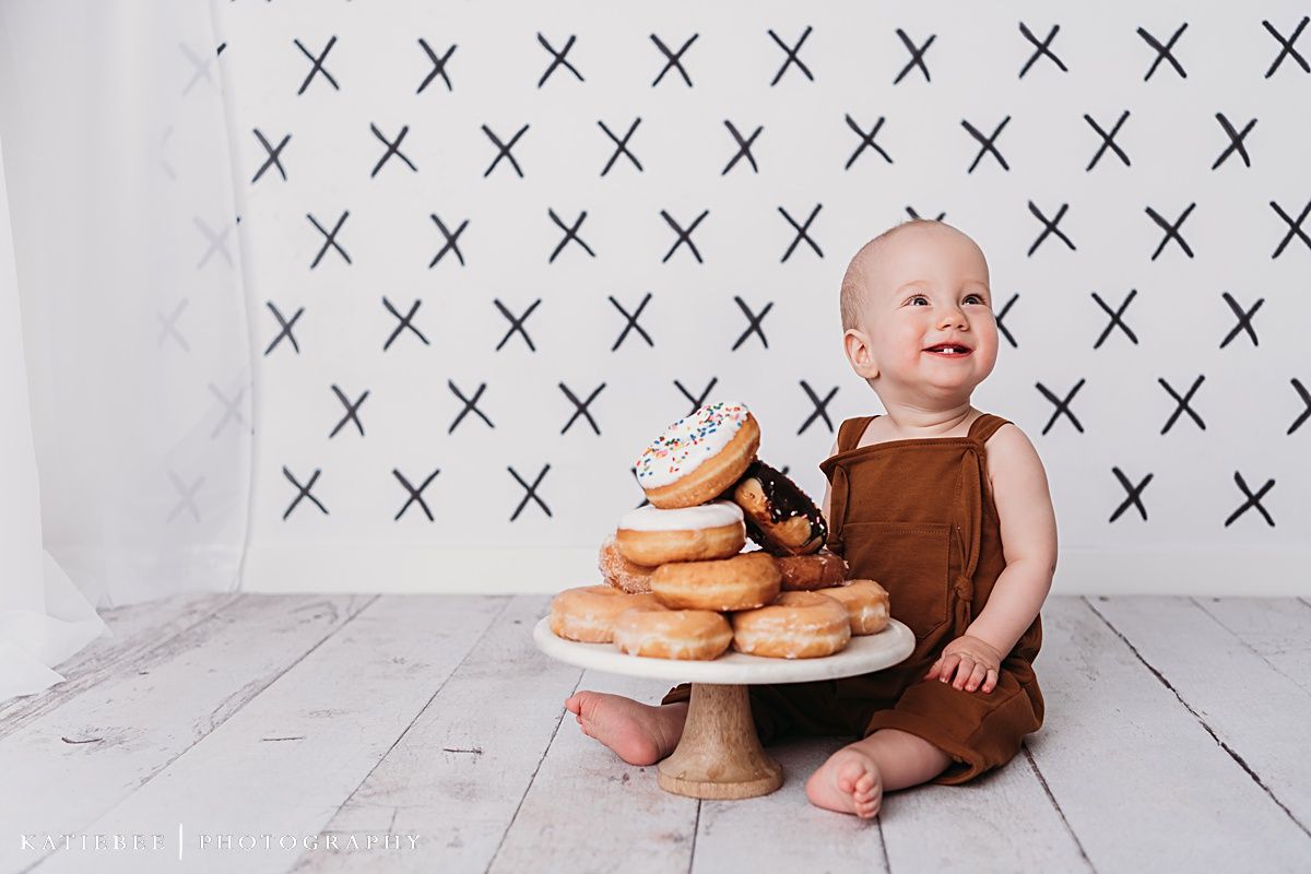 Baby sits beside elevated cake stand with stacked donuts against geometric patterned background.