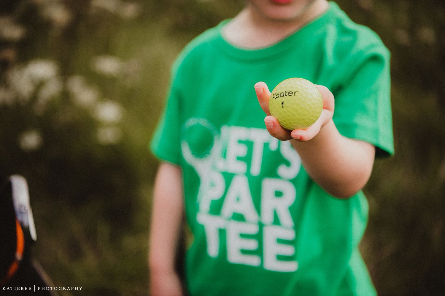 Child in green t-shirt holds tennis ball outdoors.