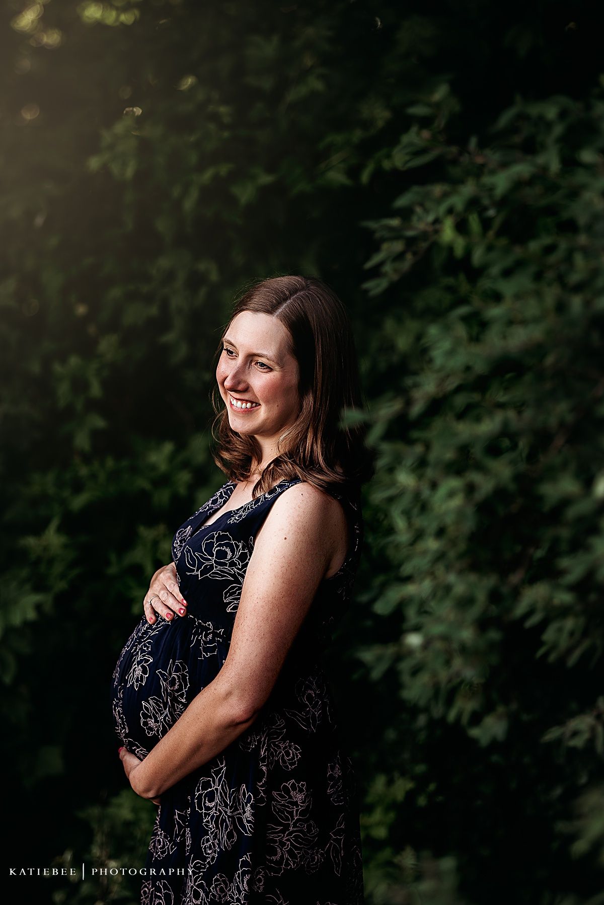 A maternity portrait featuring a woman in a navy patterned dress against green foliage.