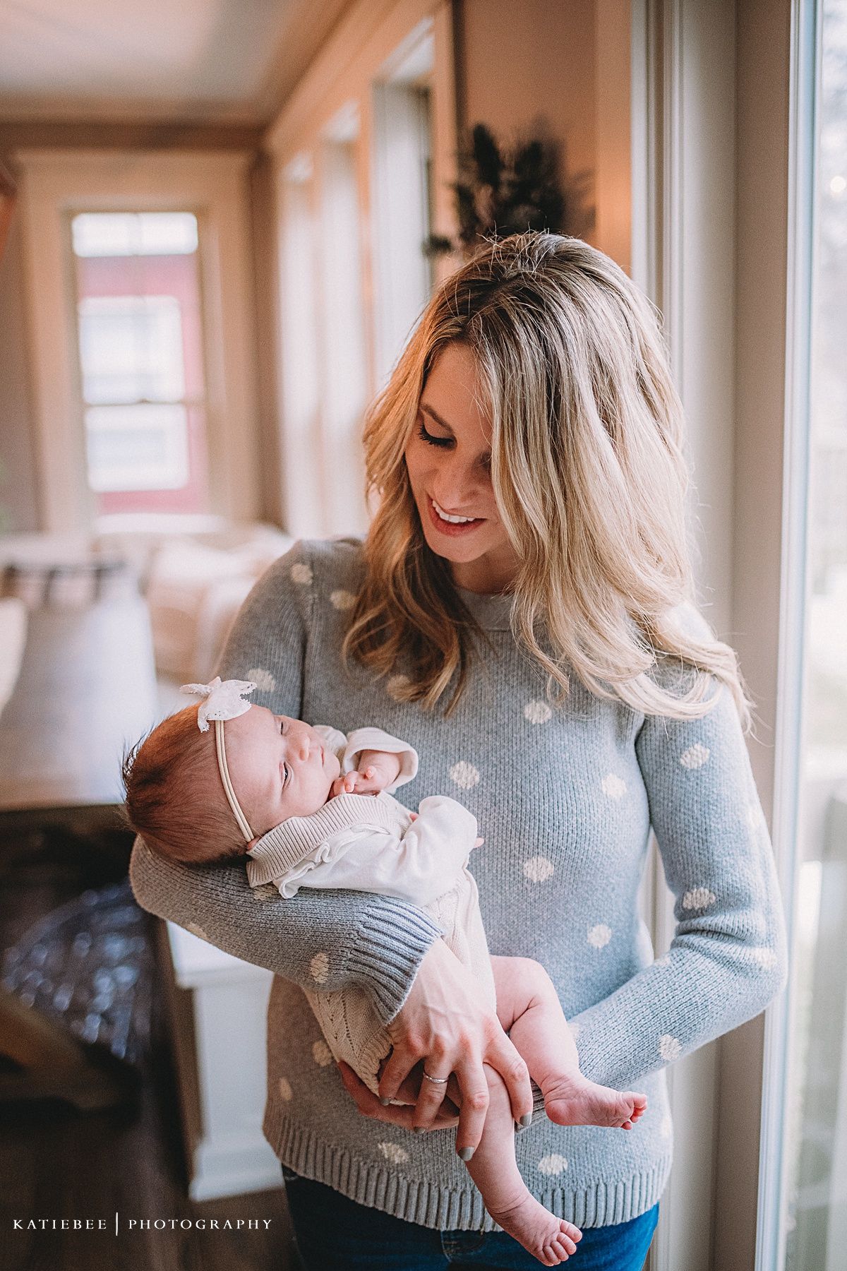 A polka dot sweater-clad figure holds a sleeping newborn baby near a sunlit window.