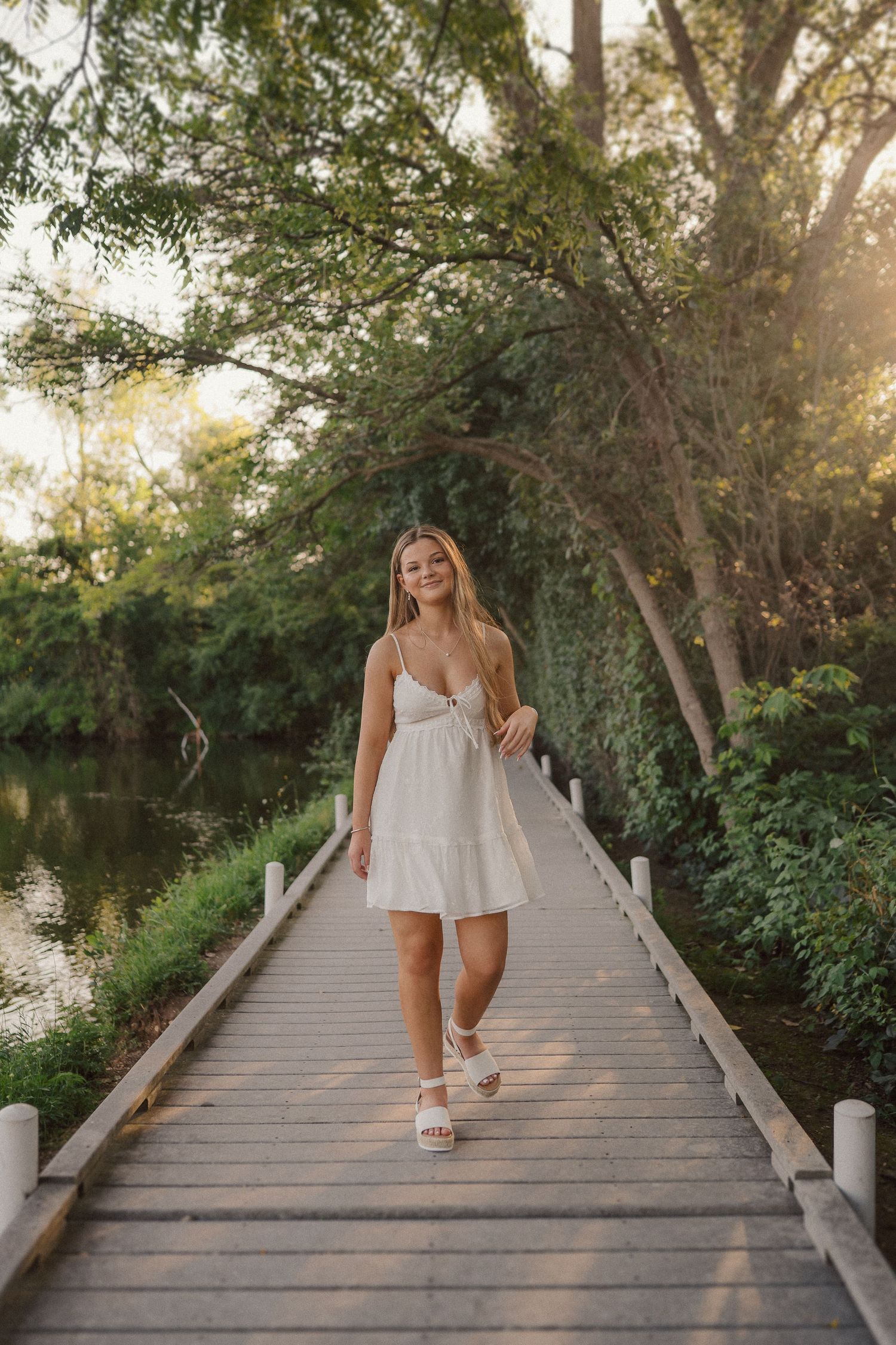 A dreamy portrait on a wooden boardwalk over water during sunset wearing a white summer dress.